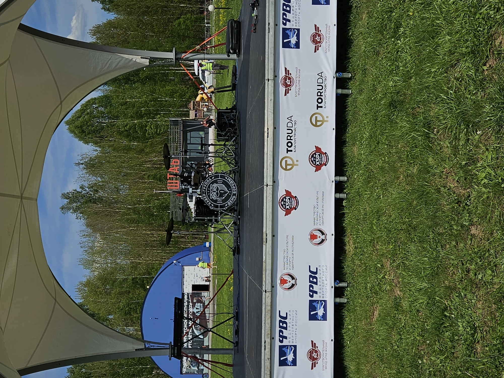 The image shows a stage set up outdoors, likely in Izhevsk, Russia, with a canopy structure partially visible on the left side. A drum kit is positioned on the stage, behind which is a backdrop or signage. The signage contains logos and text, including "PBC ФЕДЕРАЦИЯ ВЕРТОЛЕТНОГО СПОРТА РОССИИ", "МИНИСТРЕСТВО ПО ФИЗИЧЕСКОЙ КУЛЬТУРЕ И СПОРТУ УДМУРТСКОЙ РЕСПУБЛИКИ", "TORUDA БЛАГОУСТРОЙСТВО", "БЛАГОТВОРИТЕЛЬНЫЙ ФОНД ШТАБ ЖИЗНИ" and "XSPI". A blue screen is also visible.
In the background, trees are visible under a blue sky with some clouds. To the right of the stage is a patch of green grass. A person is seen on the stage in the distance.
The image is rotated 90 degrees counter-clockwise.