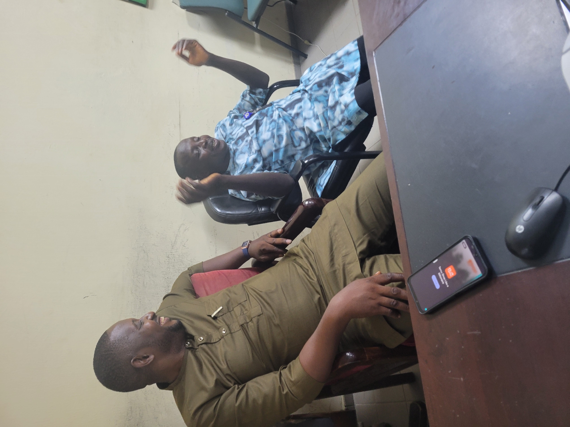 The image captures two African men indoors, presumably in an office setting, with a desk featuring a smartphone and a computer mouse. The man closer to the camera is seated in a chair and appears to be laughing while looking upwards and possibly gesturing. He is wearing a tan-colored, long-sleeved, button-down shirt or tunic. A wristwatch is visible on his left wrist. The other man is seated in a swivel chair with armrests, reclining backward, and gesturing upwards with his right hand. He is wearing a blue-and-white patterned shirt. The environment includes a beige-colored wall and dark desk. The image seems to have been taken with a slight vertical tilt, and the quality is somewhat grainy. While it is challenging to pinpoint the exact location within Abuja, Nigeria, the indoor office setting and the presence of two men suggest it is likely a workplace.