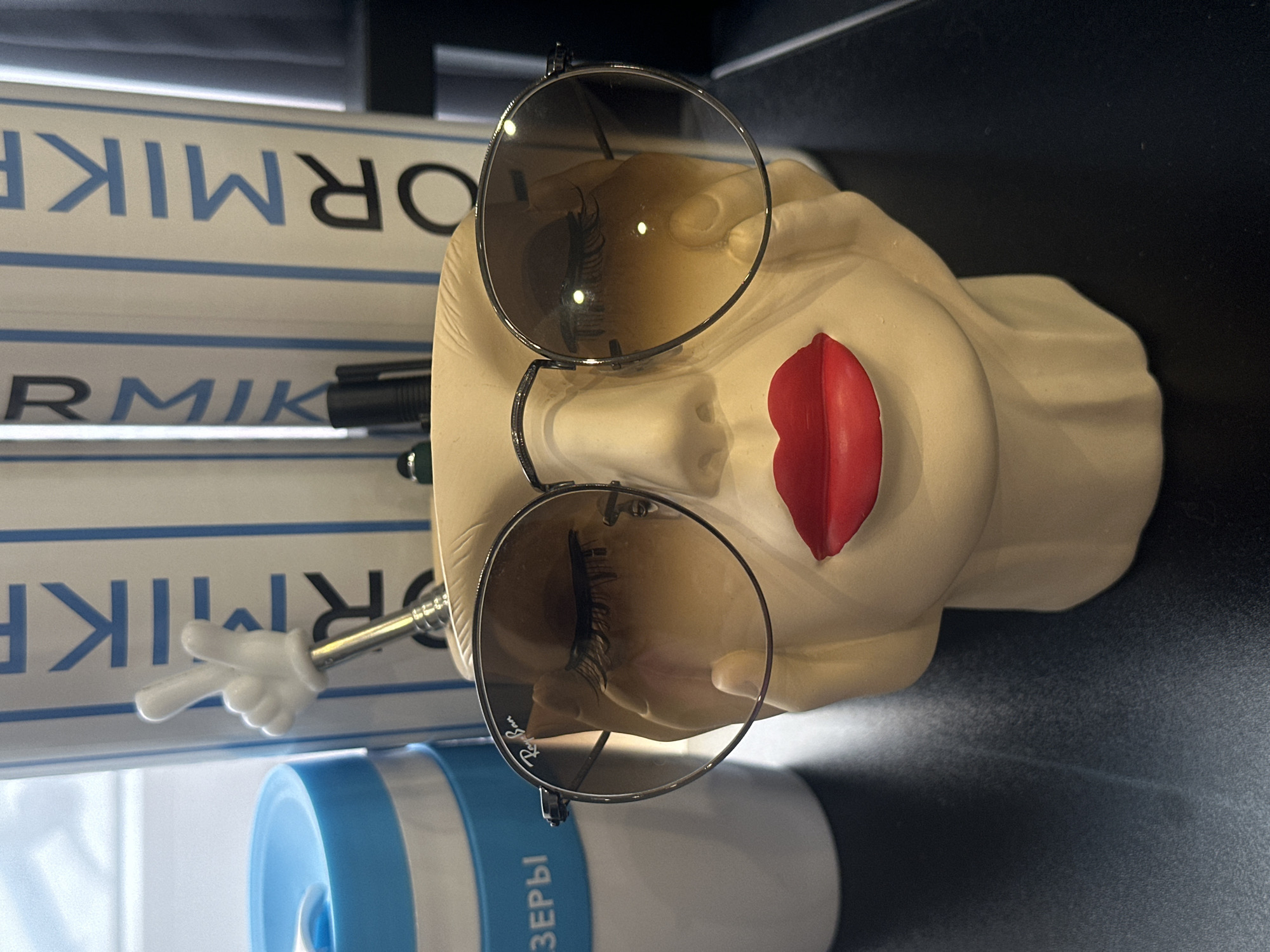 A low-angle, close-up studio shot shows a pale, possibly ceramic bust with oversized red lips and round Ray-Ban sunglasses perched on its face. The right side of the image has a dark surface, likely a tabletop or shelf. To the left of the bust is a cylindrical object with blue and white bands, labeled with the word "ЛАЗЕРЫ" (Lasers) in Cyrillic characters. Behind these objects, vertical white strips labeled with a repeated word "ОРМИК" (ORMIC) in blue, block lettering are visible. A small, white plastic hand is positioned so that it appears to be holding onto the sunglasses. The overall composition is staged and designed for product display. There is no mention of the city of Riga, Latvia in the image.