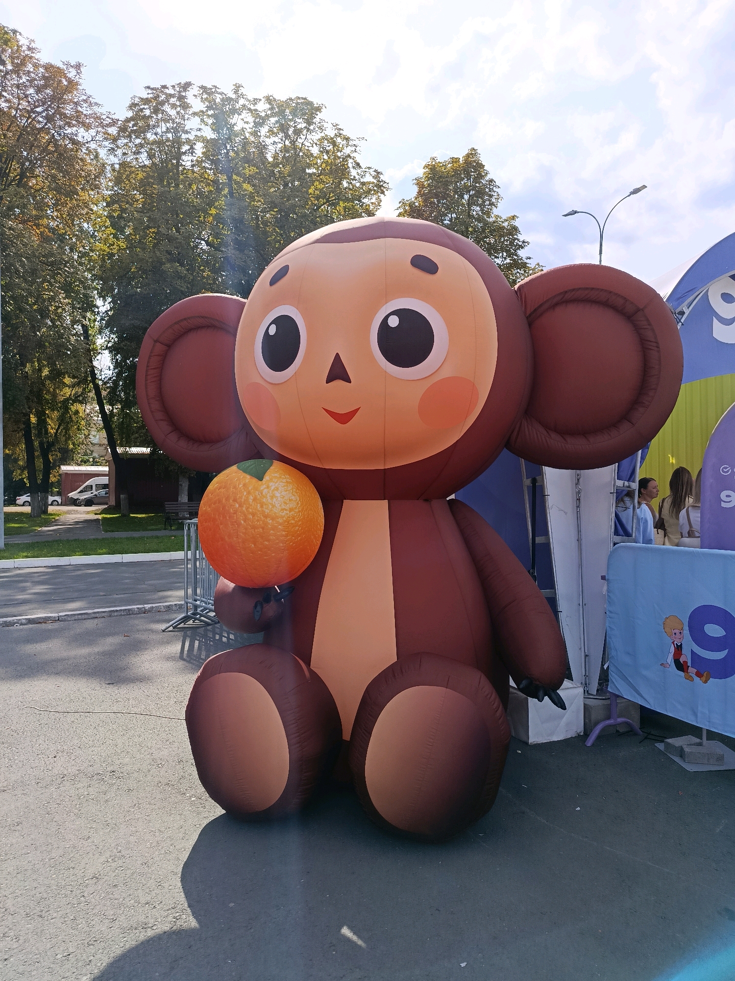 An inflatable Cheburashka character, a popular Russian cartoon character, is seated outdoors. The character is large, with brown, rounded ears and a light brown face with large black eyes and pink cheeks. It is holding a bright orange, spherical object that resembles a tangerine or clementine in its left hand. Cheburashka is positioned on asphalt, with a shadow cast to its left. Behind the character, a blue banner with a cartoon drawing of a young boy and the number "9" is visible, along with another banner that is partially obscured. Trees with green foliage are in the background, and there is a street with a white van on the left side of the image. The scene appears to be an outdoor event or festival, possibly in a city park or public square in Saratov, Russia.