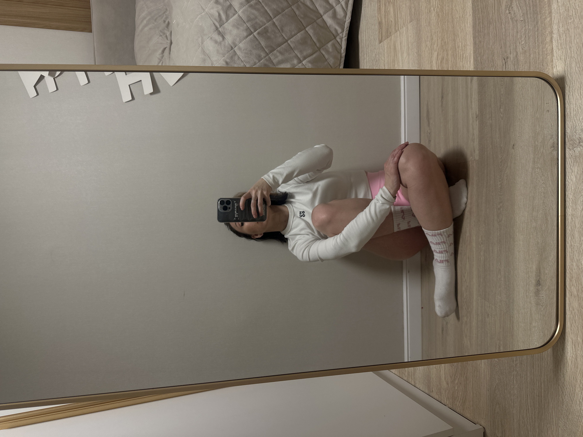 The image shows a mirror selfie of a woman wearing a white long-sleeved shirt, pink shorts, and striped socks. She is seated on the floor with her legs curled up and her face mostly obscured by the phone. The background includes a wall with white decorative elements, flooring, and a portion of a bed with a quilted comforter. There is no information available about the location in Votkinsk, Russia.