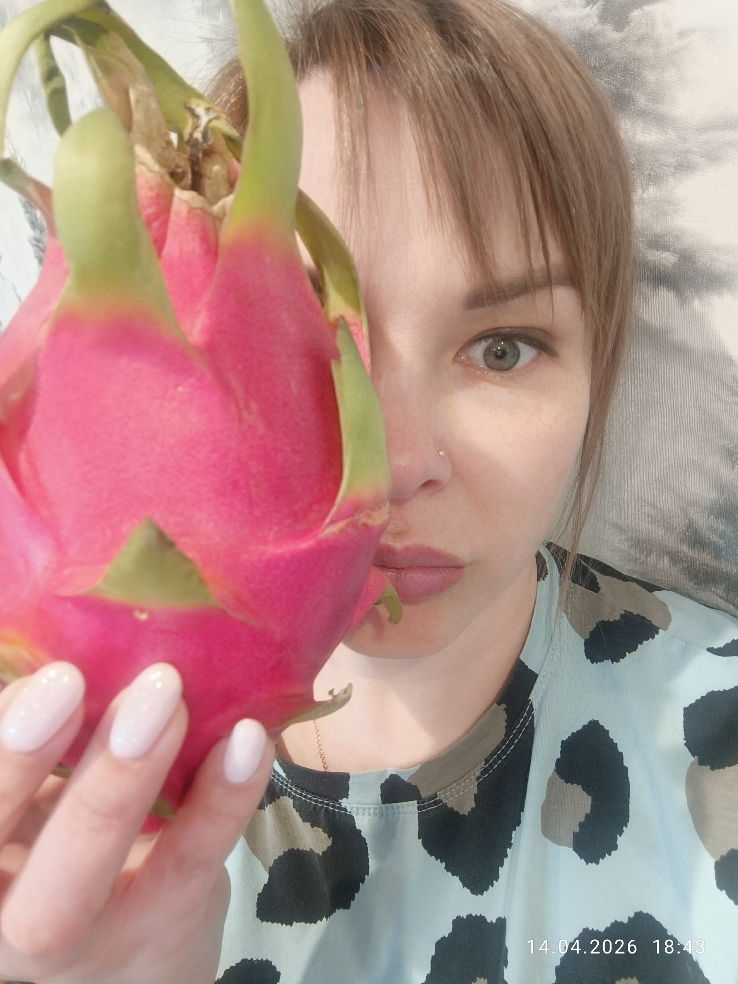 A woman is holding a dragon fruit in front of her face. She is wearing a light blue shirt with a black and brown animal print. Her fingernails are painted pale pink. She has light brown hair and grey eyes. The background appears to be a grey and white patterned wall. There is a timestamp in the lower right corner reading "14.04.2026 18:43". There is no information in the image to indicate the city is Chaikovsky, Russia.