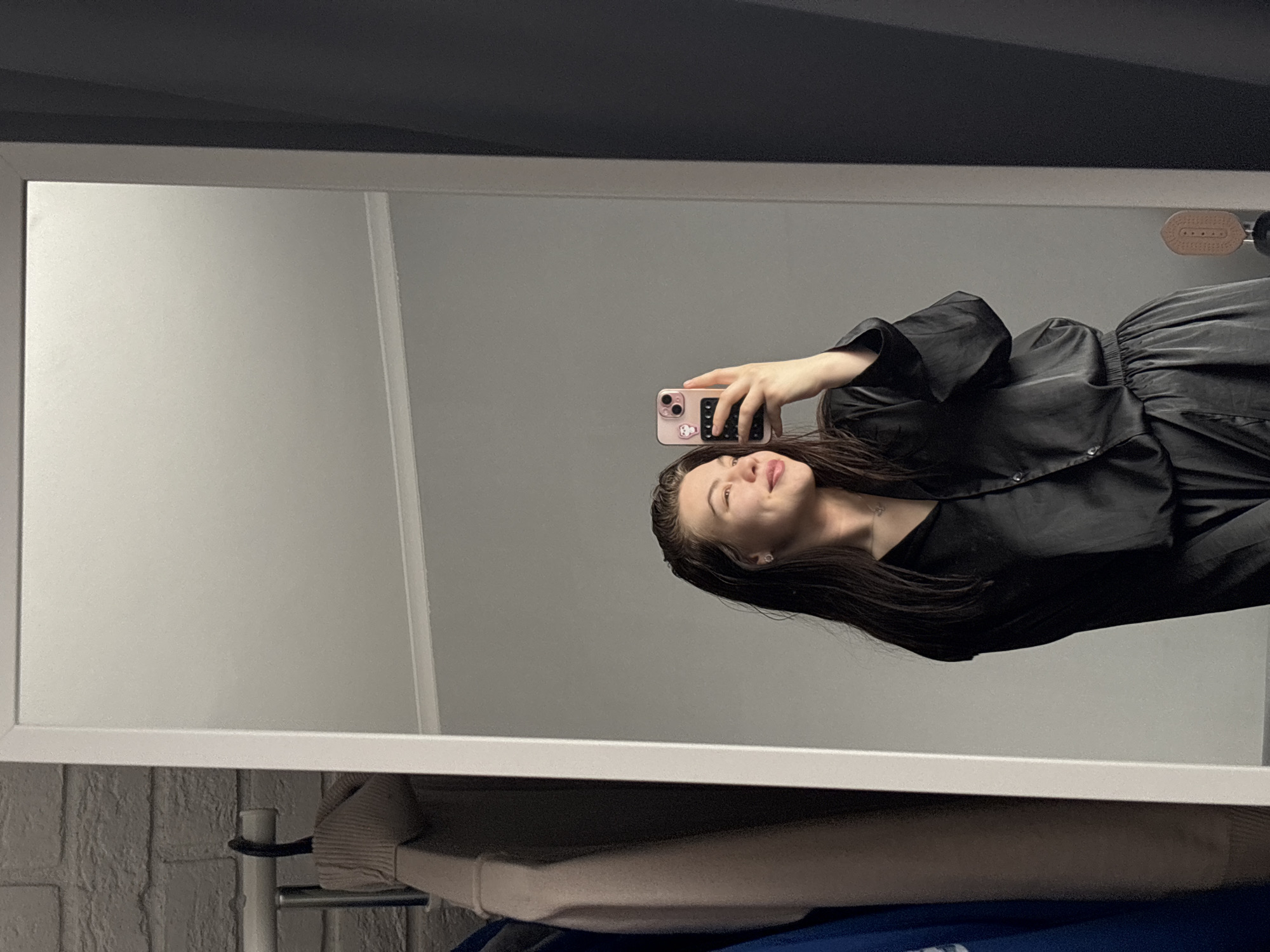 A person is taking a picture of themself in a mirror. The person has long dark hair and is wearing a black, buttoned garment. The mirror has a white frame and reflects a gray wall. Below the mirror, a patterned brick wall, a shelf with miscellaneous objects, and possibly part of a clothes rack are visible. Based on limited information and an inference, the location might be in Perm, Russia.