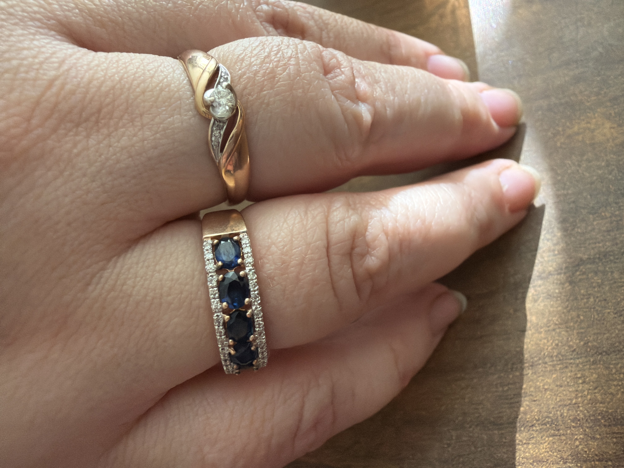 The image shows a close-up of a hand with two rings. The top ring is gold with a diamond set in the center, flanked by smaller diamonds. The bottom ring is gold with a row of alternating blue sapphires and diamonds. The hand is resting on a wooden surface with a light source from the right, creating shadows. There is no indication of Bugulma, Russia, or any other location.