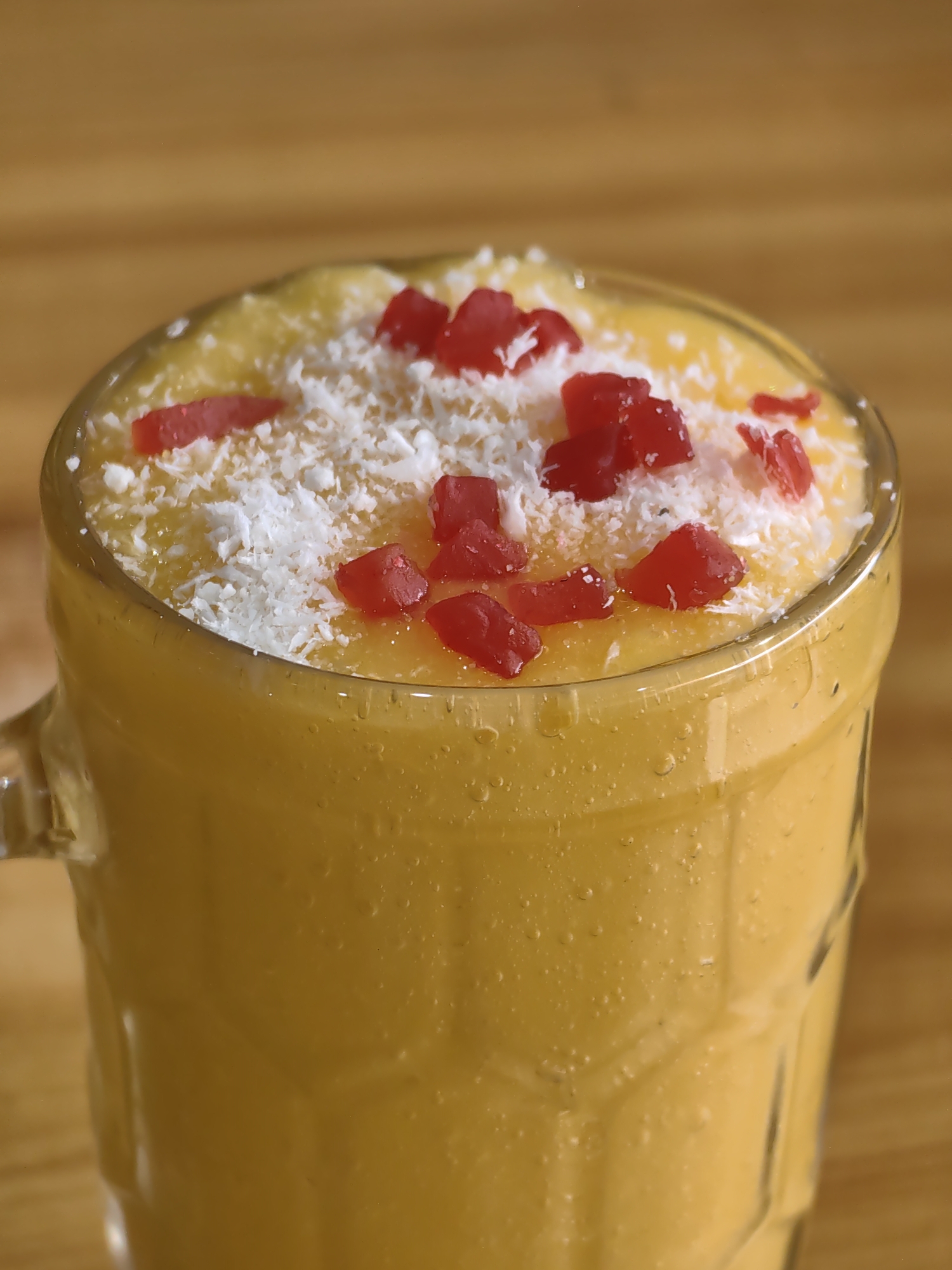 A close-up, eye-level shot shows a tall glass mug filled with a thick, orange beverage, likely a mango lassi or smoothie. The drink is topped with a sprinkle of shredded coconut and small, bright red candied fruit pieces. The glass mug has a handle on the left side and a textured surface with visible bubbles. The beverage is a vibrant yellow-orange color. The mug rests on a wooden surface, possibly a table, with a blurred wooden background. The lighting is warm and natural, highlighting the texture and color of the drink. The scene appears to be set in Vashisht, India, a town known for its hot springs and temples.