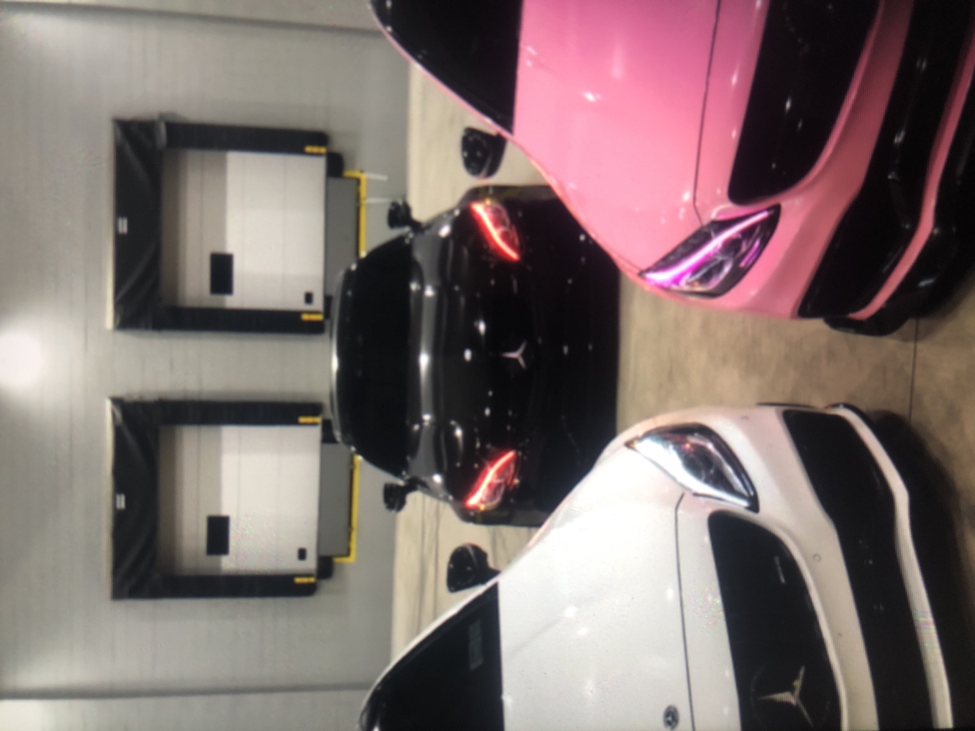 The image shows three Mercedes-Benz cars, in a warehouse setting with loading docks. The cars are: a pink and black model, a black model, and a white and black model. The black car is positioned between the white and pink vehicles. The environment is a large warehouse or storage area, as evidenced by the presence of two loading dock doors. I am unable to determine the location in the city of Lhokseumawe, Indonesia.