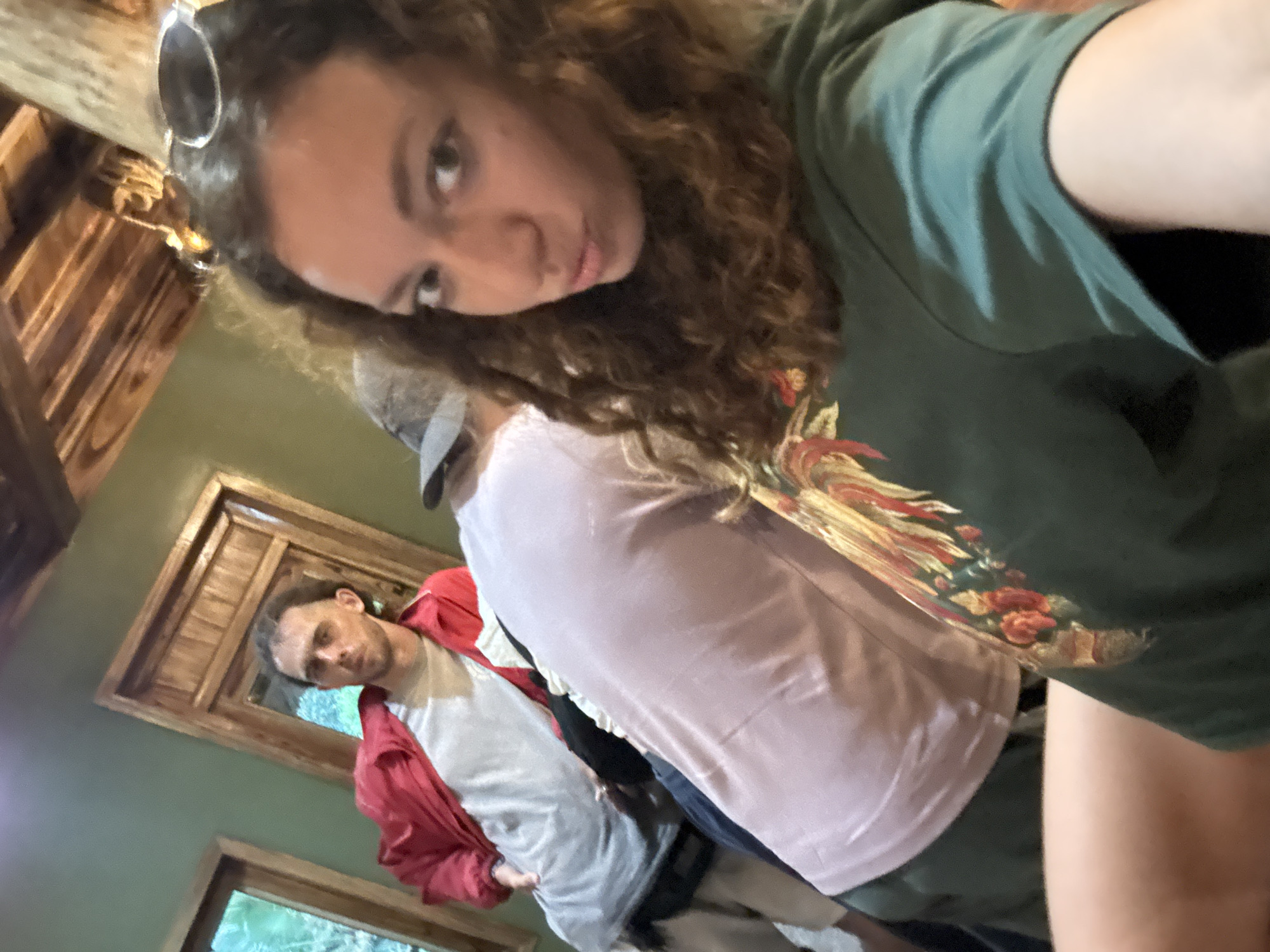 In a rustic, wooden-beamed interior, likely within Pù Luông Commune, Vietnam, a group of individuals are captured in a candid moment. A young woman with curly brown hair, wearing a green hooded sweatshirt with a floral design, takes a selfie, puckering her lips at the camera. Behind her, a man in a red jacket and a light-colored shirt is partially visible, his gaze directed away from the camera. Another person, wearing a light pink t-shirt, is also in the background, their back to the viewer. The setting suggests a casual gathering or a moment of rest in a communal area, with warm wooden tones and a muted green wall contributing to the atmosphere. Natural light streams in from windows, highlighting the textures of the wooden ceiling and furniture.