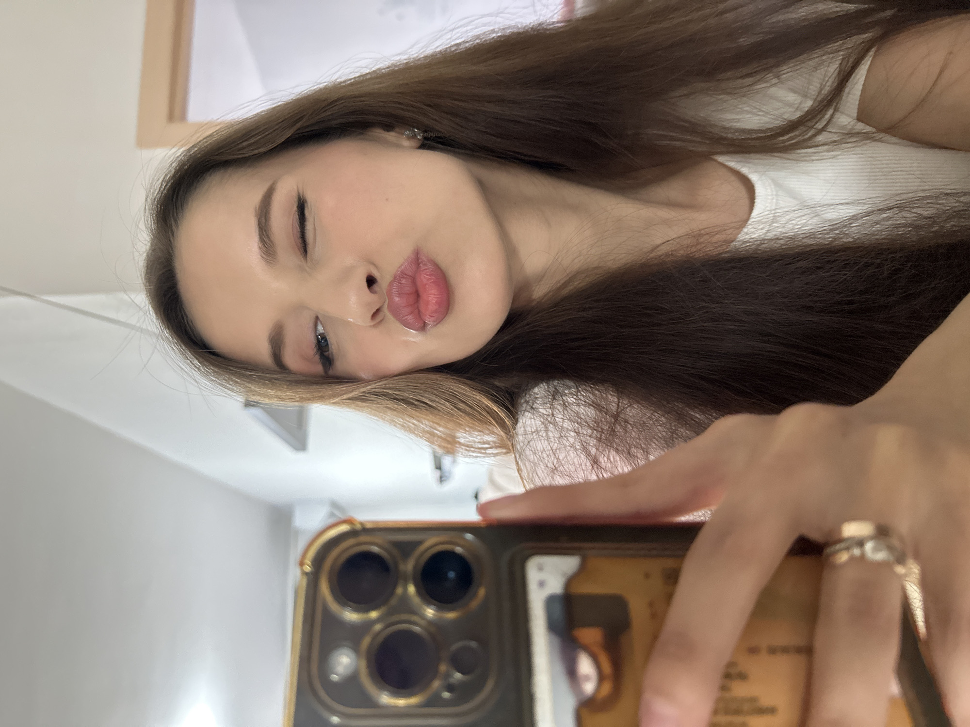 A selfie of a young woman with long, brown hair. She has fair skin, light eye makeup, and pink lipstick, pursing her lips slightly toward the camera. She is wearing a white top. The woman is holding a smartphone in front of her, partially obscuring the view. The phone case appears to be brown and has multiple camera lenses visible. The background is a simple, neutral-toned wall with a frame visible in the upper left corner. The setting appears to be indoors, likely in a private residence. It is not possible to identify the location in Yekaterinburg, Russia from the image.