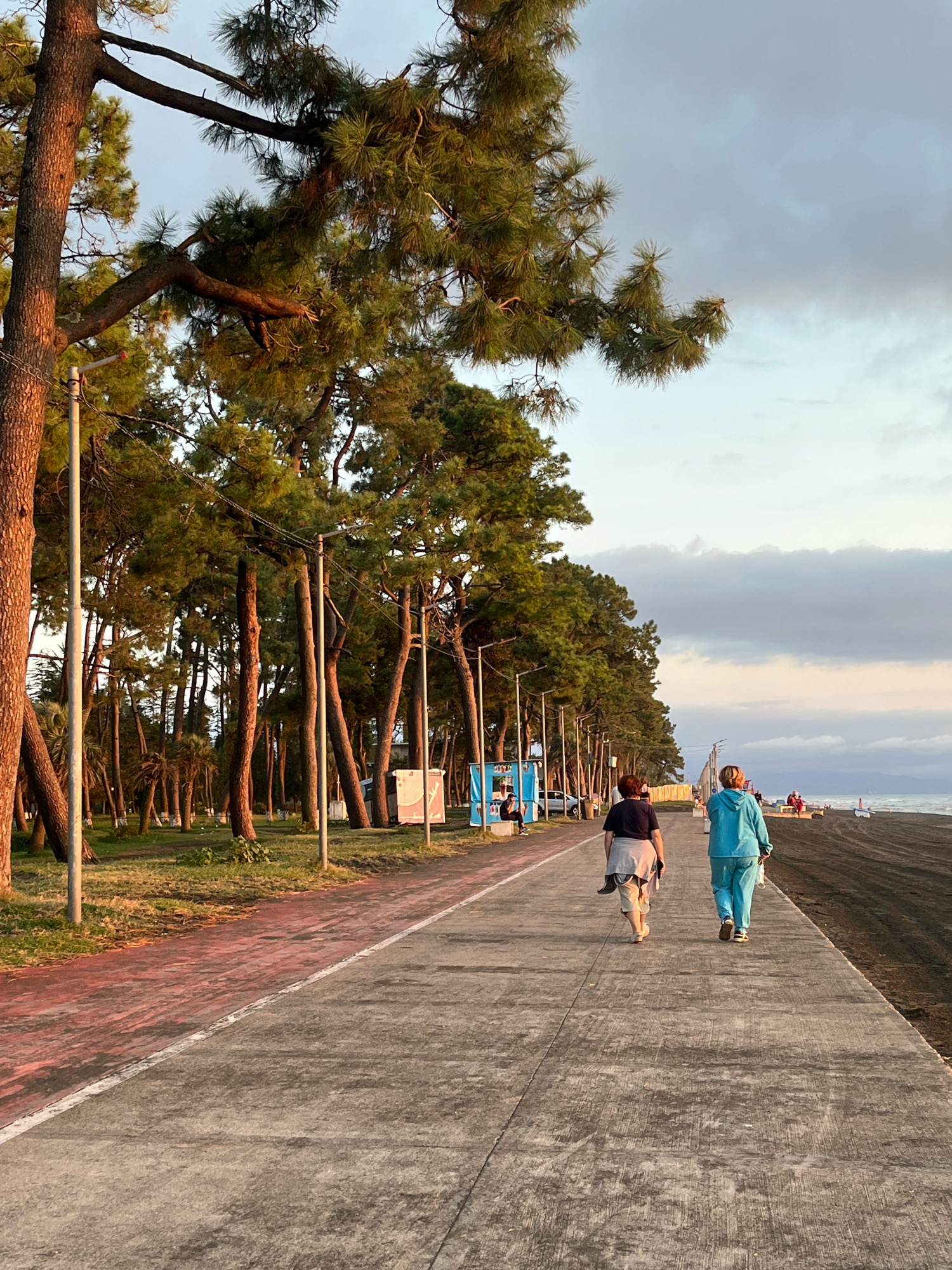 The image shows a paved walkway along a beach in Ureki, Georgia. Two women are walking away from the camera on the walkway. The woman on the left is wearing a dark shirt and light-colored pants, and a gray cardigan draped over her shoulders. The woman on the right is wearing a blue hooded sweatshirt and blue pants. Behind them, a line of tall pine trees borders a grassy area. Further down the walkway, there are a few more people and a parked car. The beach to the right is dark sand, and the sea is visible in the distance under a cloudy sky. Streetlights are positioned along the walkway.