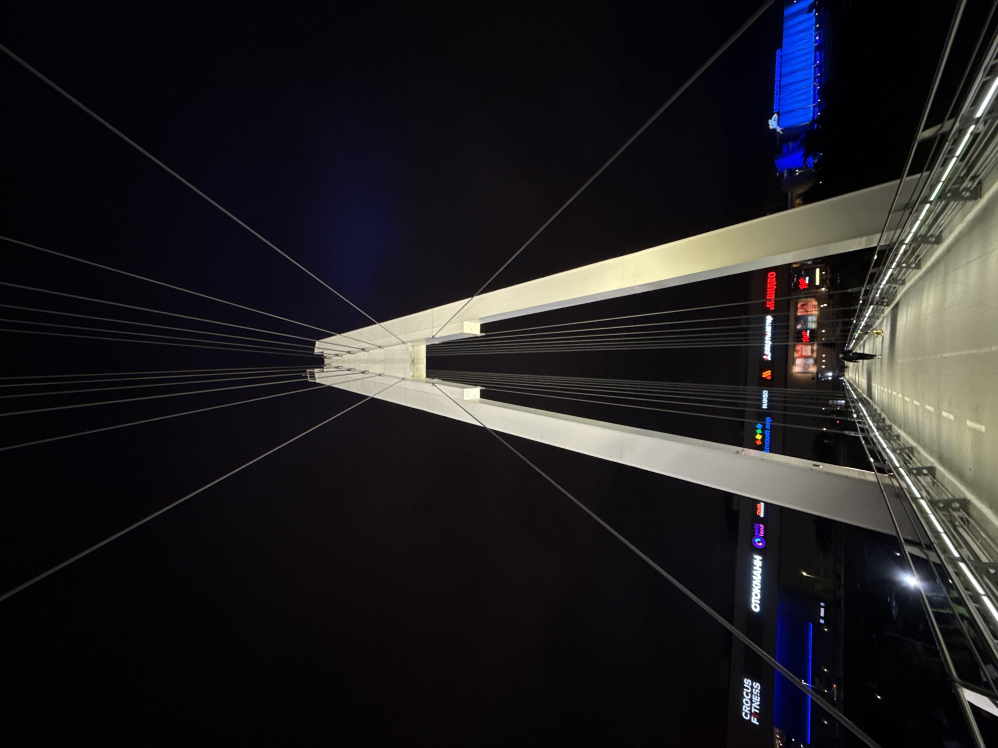 The image depicts the pedestrian bridge connecting the Crocus City Hall and Crocus Expo exhibition center in Khimki, Russia, at night. The bridge features a prominent white pylon from which cables extend upwards to support the structure. Illuminated signs of various businesses, including "CROCUS FITNESS," "СТОКМАНН," "land," "ДЕТСКИЙ МИР," and "MANGO," are visible on the buildings lining the walkway. A solitary figure, presumably a person walking a dog, is captured in the mid-ground on the bridge. The surrounding environment is dark, with the primary illumination coming from the artificial lights of the buildings and the bridge itself.
