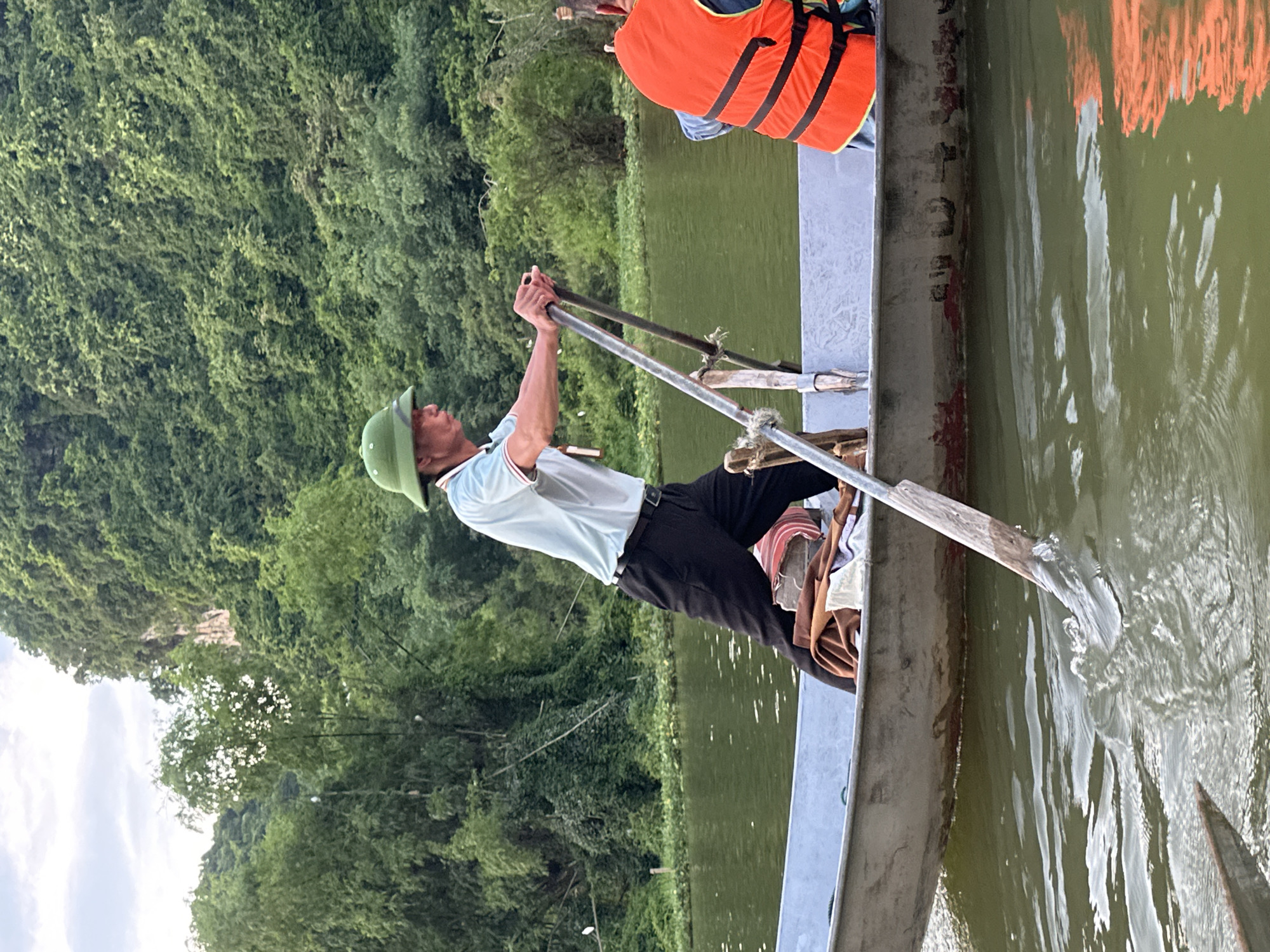 A man in a light blue polo shirt, black pants, and a green hat rows a flat-bottomed boat on a calm, green body of water. He holds a long oar with a wooden handle and a metal blade. Beside him, another person wearing an orange life vest is visible. The boat is made of metal and shows signs of wear. In the background, a lush, green mountainside covered in dense vegetation rises steeply. The water reflects the overcast sky and the surrounding foliage. The scene appears to be in a natural, possibly rural setting in Yên Sơn Ward, Vietnam, characterized by its mountainous landscape and waterways.