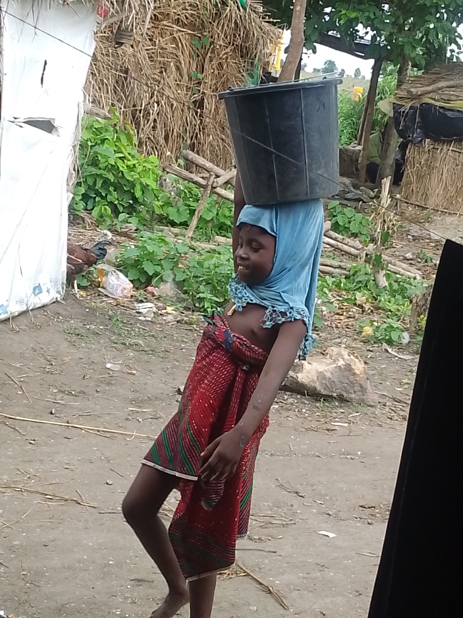 A young girl, likely of Igbo descent, walks outdoors in Otulu, Nigeria, carrying a large black bucket on her head. She is dressed in a red and green patterned cloth skirt and wears a blue headscarf that drapes over her shoulders. The ground is dirt and littered with debris. In the background, a white tarp-covered structure stands to the left, while thatched-roof huts and vegetation are visible to the right, suggesting a rural or semi-rural setting. The overall scene depicts a common activity of carrying water in a community.
