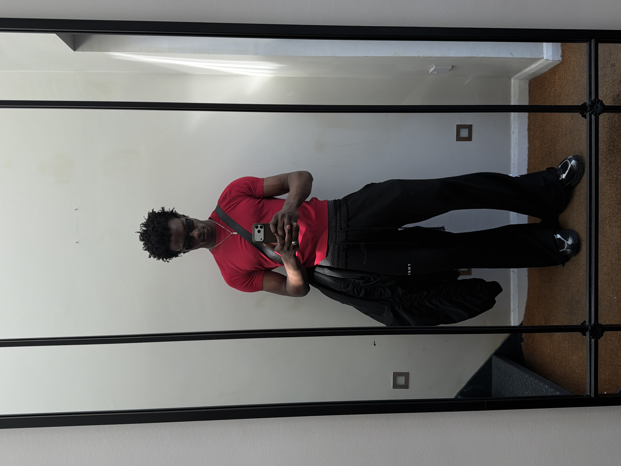 A black man is standing in front of a large, rectangular mirror in what appears to be an apartment hallway. He is wearing a red short-sleeve shirt, black sweatpants, and black shoes with white detailing. He is also wearing a silver chain, sunglasses and has a black shoulder bag across his chest. He is holding a phone, presumably taking the picture. The background shows a white wall and a hallway with tan/brown carpeting. There are electrical outlets visible on the wall. The location is likely in an apartment building in London, United Kingdom, suggested by architectural details of the building.