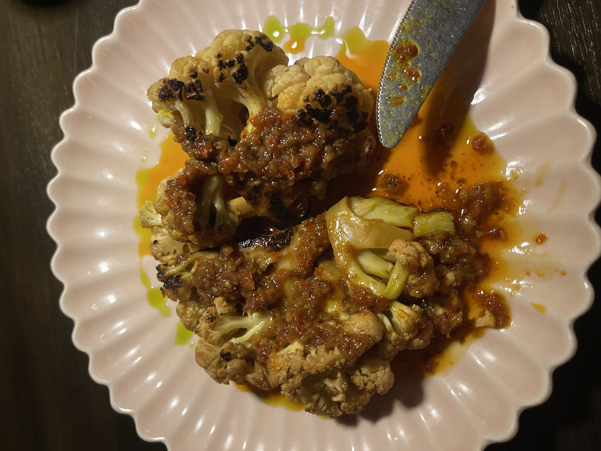 This media file depicts a close-up, overhead shot of a plate of food. The subjects are pieces of roasted cauliflower, generously coated in a rich, orange-red sauce with visible bits of chopped ingredients. The cauliflower florets show charring on the edges, indicating they were roasted. They are arranged on a light pink, scalloped-edge plate. A silver knife with a serrated edge rests on the plate, partially in the sauce, suggesting the food is being served or has been partially eaten. Small droplets of a yellowish-green sauce are also visible on the plate, adding a contrasting color to the dish. The background is dark and out of focus, hinting at a dimly lit restaurant or dining setting. The overall impression is one of a savory, potentially spicy dish served in an attractive presentation. The time of day is not discernible, and there are no specific weather cues or emotional indicators. No text is visible in the image. The location context is Mengwi, Indonesia.