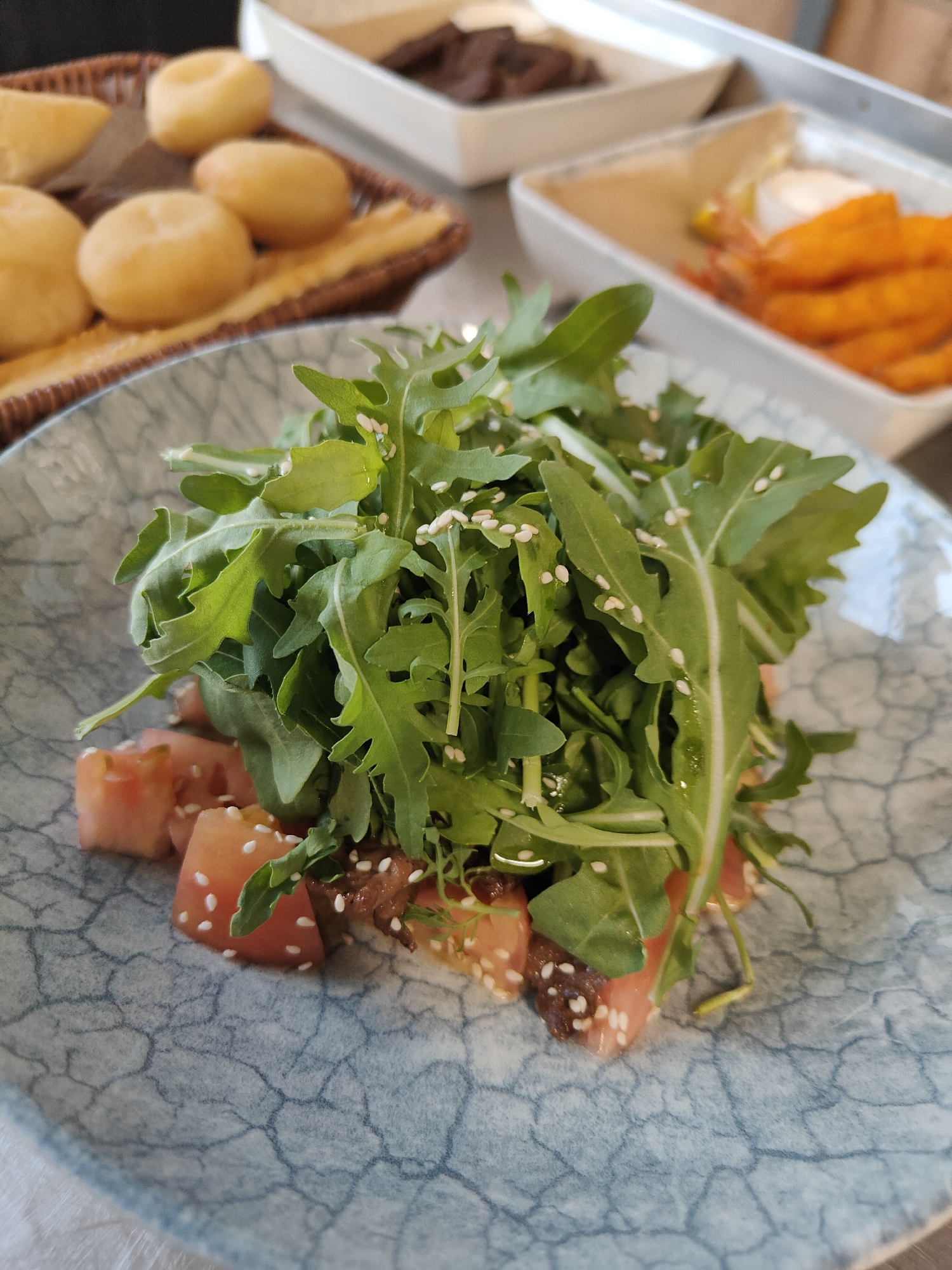 Arugula salad with diced tomatoes and sesame seeds is served on a blue patterned plate. In the background, there is a basket of bread rolls, and two white rectangular dishes containing what appear to be small pieces of meat and shrimp. The scene is indoors, possibly in a restaurant in Aktau, Kazakhstan.