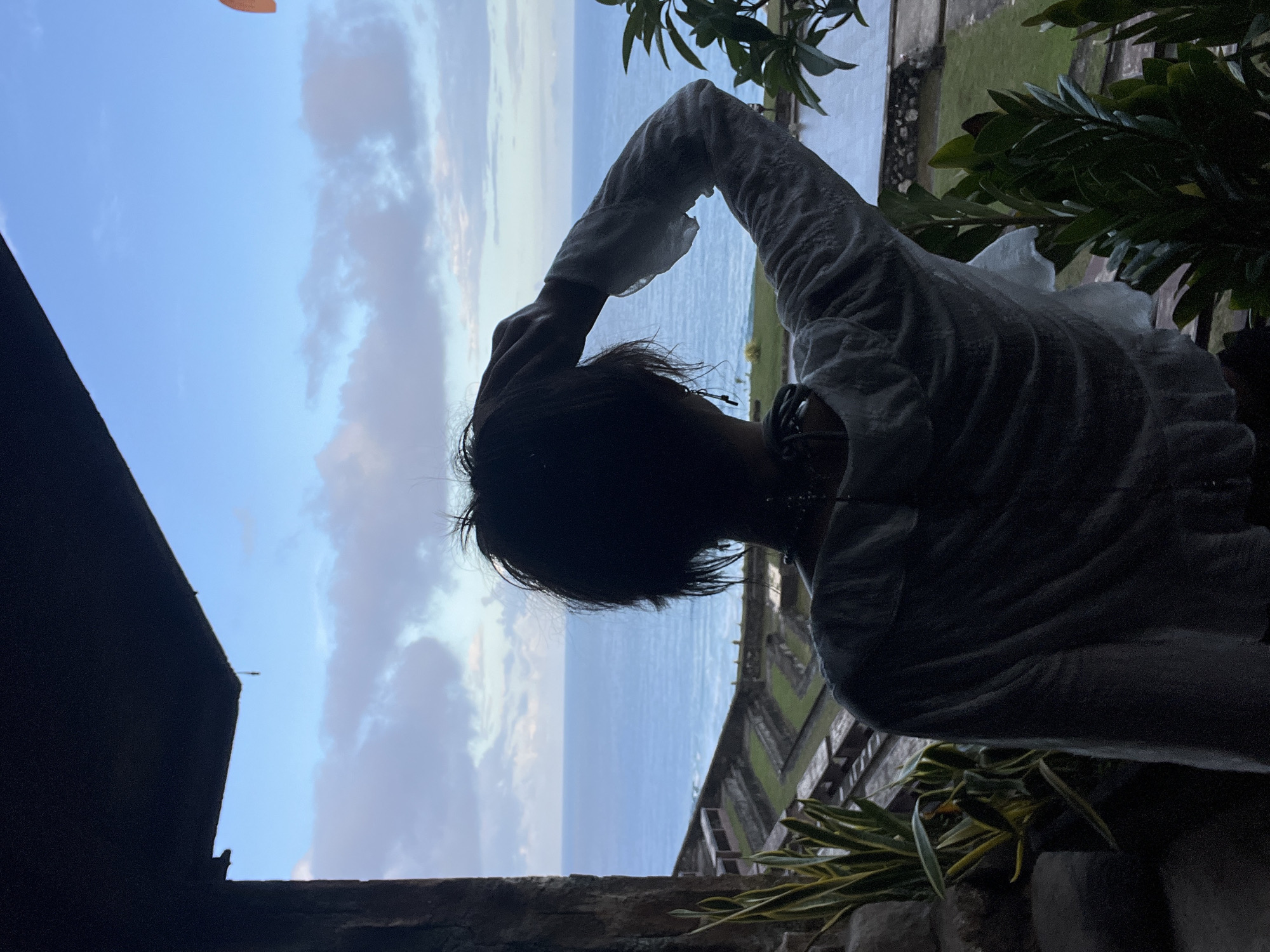 The image is oriented vertically (rotated 90 degrees counter-clockwise), showing a person in silhouette against a bright sky and ocean backdrop.
**Subjects**: The primary subject is a person, seen from behind and slightly to the side, positioned on the right half of the frame. They have short, dark hair and are wearing a light-colored, long-sleeved garment, possibly a shirt or light jacket, which appears textured or wrinkled. Their left arm is raised above their head. The background features a wide expanse of light blue sky with scattered clouds, some white and some with a light grey-pink hue, suggesting either sunrise or sunset. Below the sky is a calm, darker blue ocean stretching to the horizon. On the lower and far right edges of the frame, dark green tropical foliage and parts of a man-made structure (possibly a stone wall or railing) are visible. A dark, angular architectural element, likely a building's corner or roofline, is silhouetted on the far left.
**Activity**: The person appears to be gazing out at the ocean view, potentially taking in the scenery or shielding their eyes from the light.
**Notable details**: The image captures a scenic coastal setting, likely from an elevated viewpoint such as a balcony or terrace, overlooking the sea. The backlighting creates a strong silhouette of the person and the immediate foreground elements. The presence of tropical plants and the ocean aligns with the location context of Kuta Selatan, Indonesia. There is no visible text.