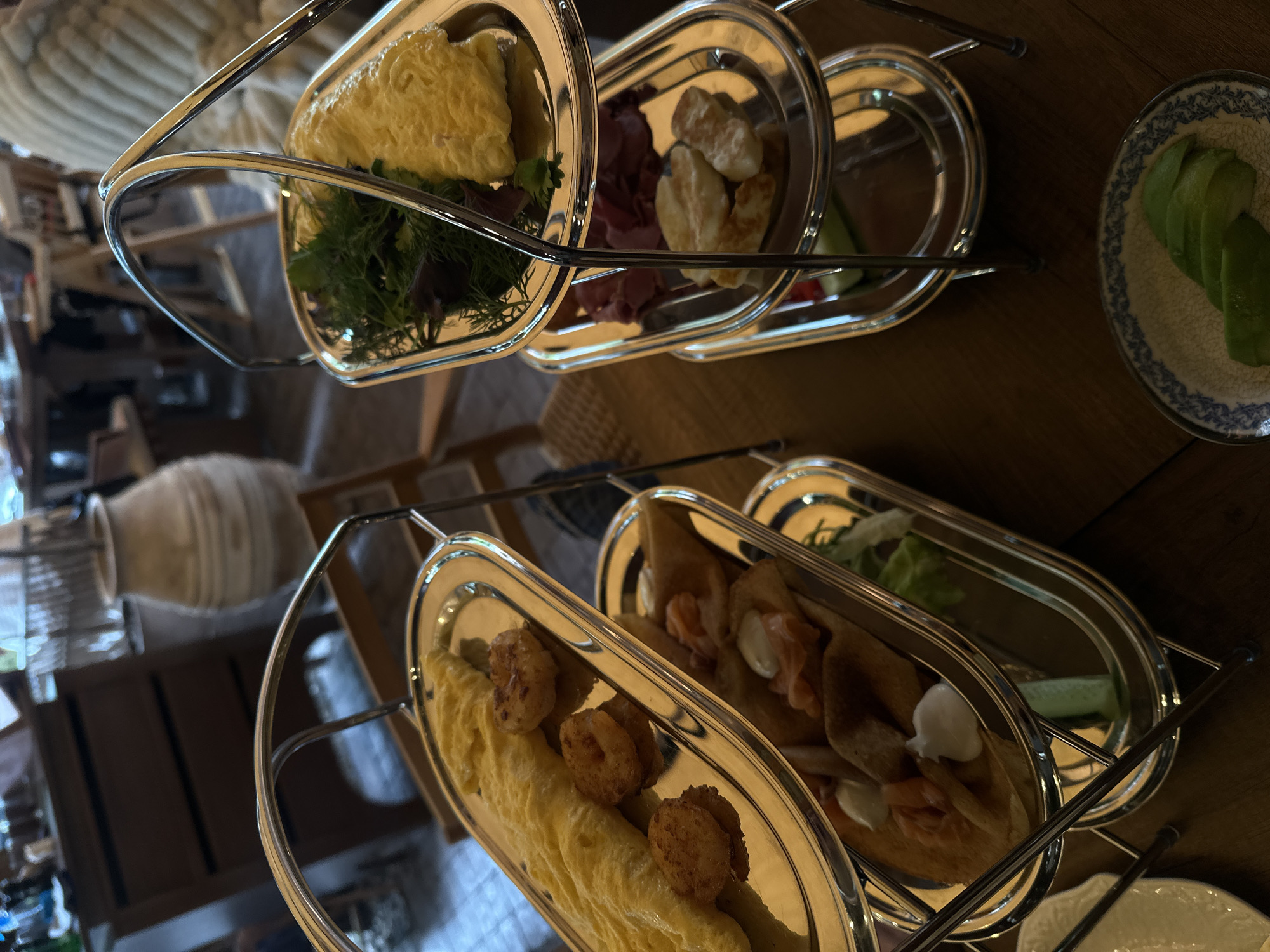The image shows a three-tiered stand of silver serving trays filled with food, likely taken in a restaurant setting. From top to bottom, the trays contain: a portion of omelet with greens; meats and fried potatoes, with cucumber slices; and pancakes with seafood and vegetables. To the right of the bottom tier, there is a plate of sliced avocados. The stand is positioned on a wooden table. In the background, out of focus, there is a light-colored decorative vase on a table, and hints of other patrons. This might be a hotel, located in Yerevan, Armenia.