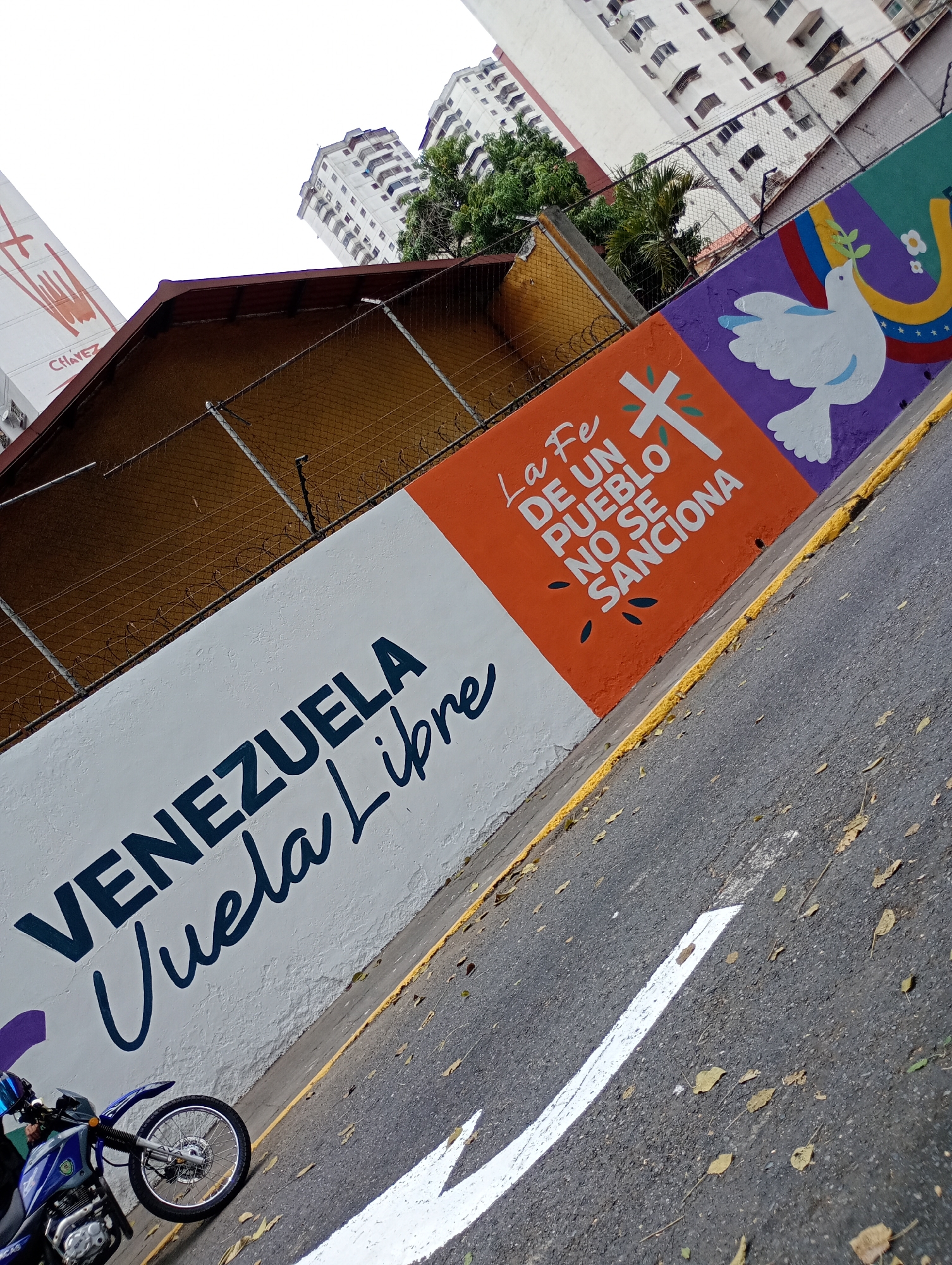 The image captures a mural on a wall in Caracas, Venezuela. The mural features the phrase "Venezuela Vuela Libre" (Venezuela Flies Free) in dark blue, followed by an orange square with "La Fe de un Pueblo No se Sanciona" (The Faith of a People Should Not Be Sanctioned) written in white around a cross. To the right of this is a purple square with a white dove holding an olive branch, followed by what appears to be parts of the Venezuelan flag. The wall is topped with a chain-link fence, and the background includes buildings and trees. A motorcycle is partially visible in the bottom left corner. The photo is taken at an angle, showing the road with a white arrow marking. The street is littered with leaves.