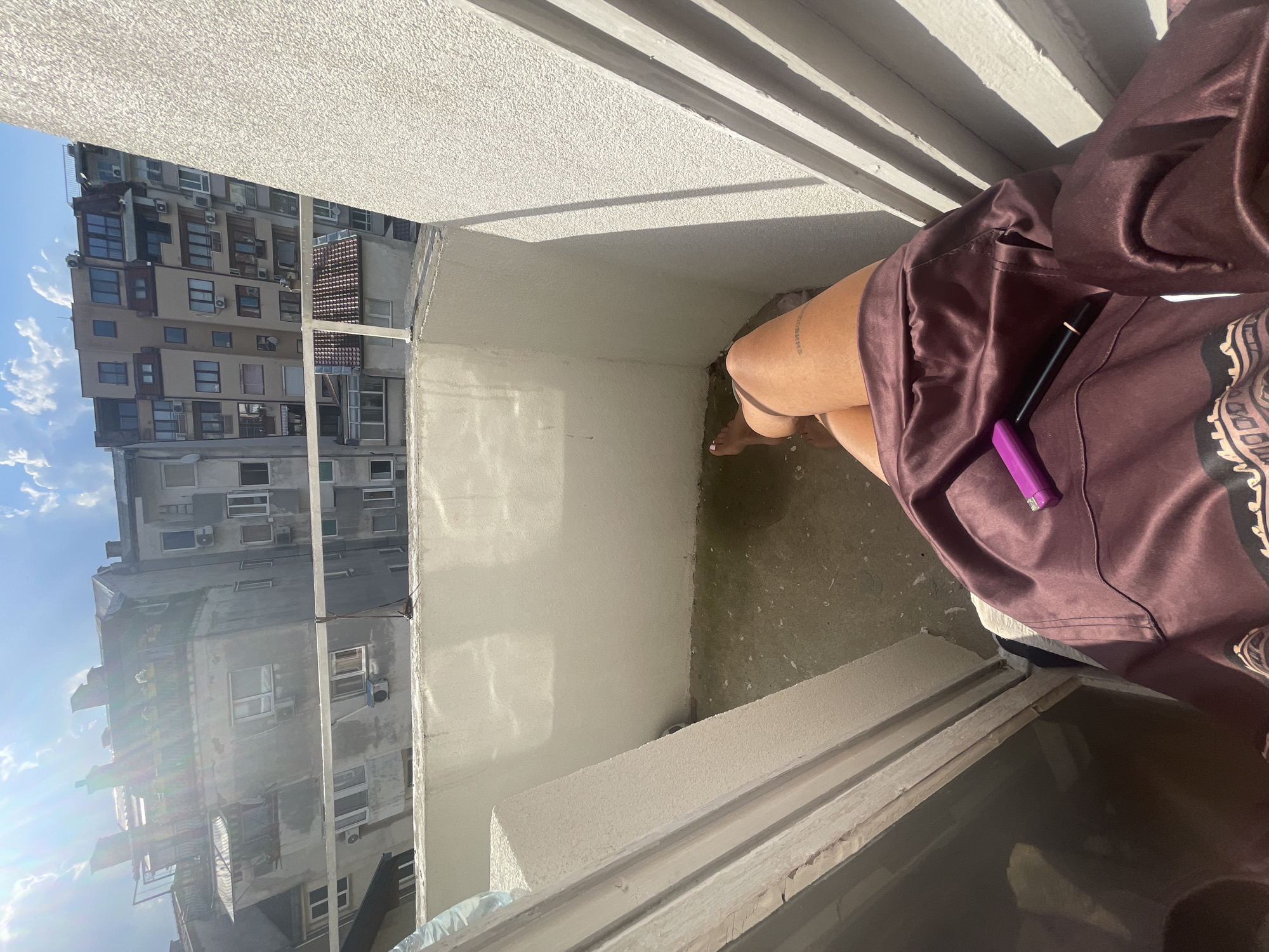 The image shows a person sitting on a balcony in an urban setting, likely Varna, Bulgaria, given the architectural style of the surrounding buildings. The person is wearing a silky, maroon robe and has a tattoo on their left thigh. A purple device and a black pen are resting on the robe. The balcony floor is dirty and appears to have some water or liquid pooled on it. In the background, several multi-story buildings with balconies, windows, and air conditioning units are visible under a bright, partly cloudy sky. The overall impression is a casual moment on a balcony on a sunny day in a dense city.