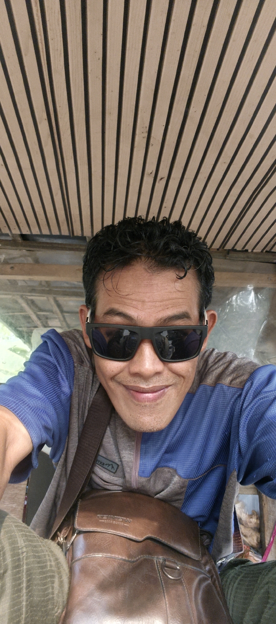 A man wearing sunglasses and a blue and gray jacket is photographed from the chest up. He is smiling and looking at the camera. The background appears to be a wooden slatted ceiling. A brown leather bag is visible across his chest. The image is likely a selfie taken outdoors in a semi-rural or casual setting, possibly near Muaro Jambi, Indonesia, given the lighting and natural elements.