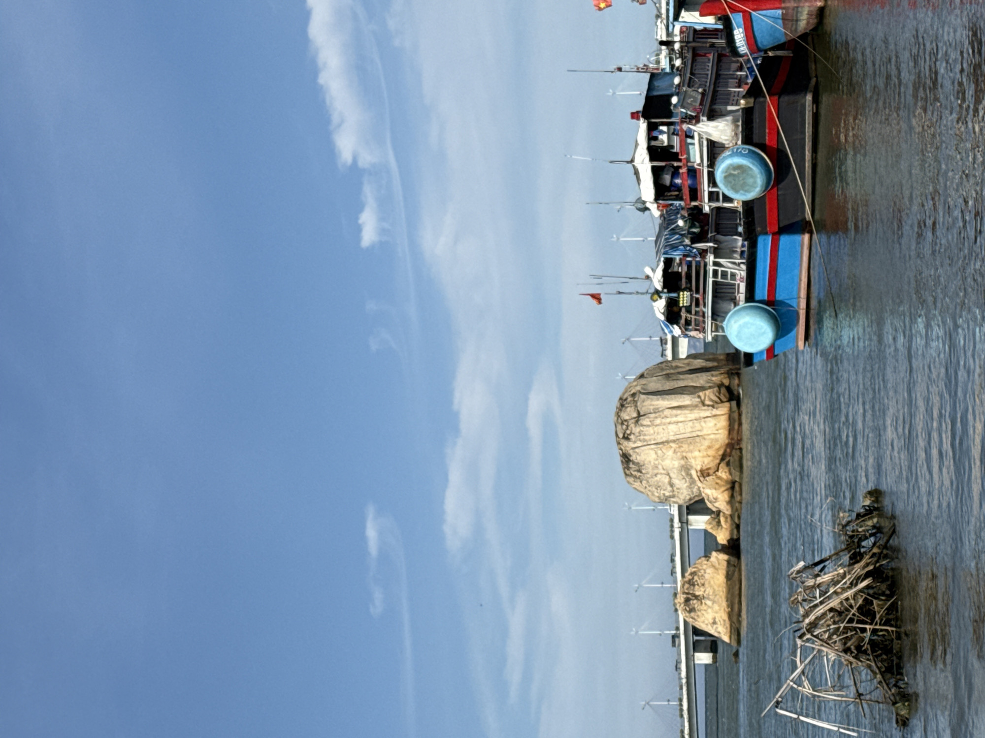 The image depicts a coastal scene in Bac Nha Trang Ward, Vietnam, under a bright blue sky with scattered white clouds. In the foreground, the calm, dark blue water is disturbed by ripples and reflections. A pile of dry branches and debris is partially submerged on the left side of the frame. To the right, two fishing boats are moored side-by-side. The boat in the foreground is painted with a blue hull and a red stripe, featuring two large blue buoys. The boat behind it has a similar color scheme with a dark hull and a red stripe. Both boats have white superstructures and appear to be equipped for fishing. In the middle ground, two large, weathered rock formations rise from the water. Further in the background, a long, modern bridge spans the water, supported by numerous pylons, with wind turbines visible along its length. The overall atmosphere is peaceful and scenic, highlighting the intersection of natural beauty and human activity in this Vietnamese coastal area.