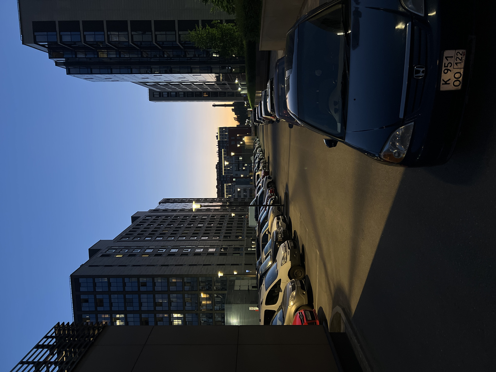 The image depicts an urban scene in Saint Petersburg, Russia, at twilight. Modern, multi-story residential buildings with dark facades and numerous glass windows line both sides of a paved street or parking area. Several internal lights are visible within the building windows. Artificial streetlights, emitting a warm yellow glow, are active along the thoroughfare, casting long shadows on the asphalt. The sky exhibits a deep blue overhead, transitioning to an orange and yellow hue at the horizon. In the distance, a tall, slender industrial chimney or tower is silhouetted against the twilight sky. Numerous cars are parked in rows along the street. In the immediate foreground, a dark blue Honda sedan is prominently featured, displaying a Russian license plate with the registration "K 951 OO 122 RUS". Other visible vehicles include a white panel van, a gold-colored sedan, and various other dark-colored cars. Landscaping with bushes and small trees is present along the base of the buildings on the right. No individuals are present in the scene.