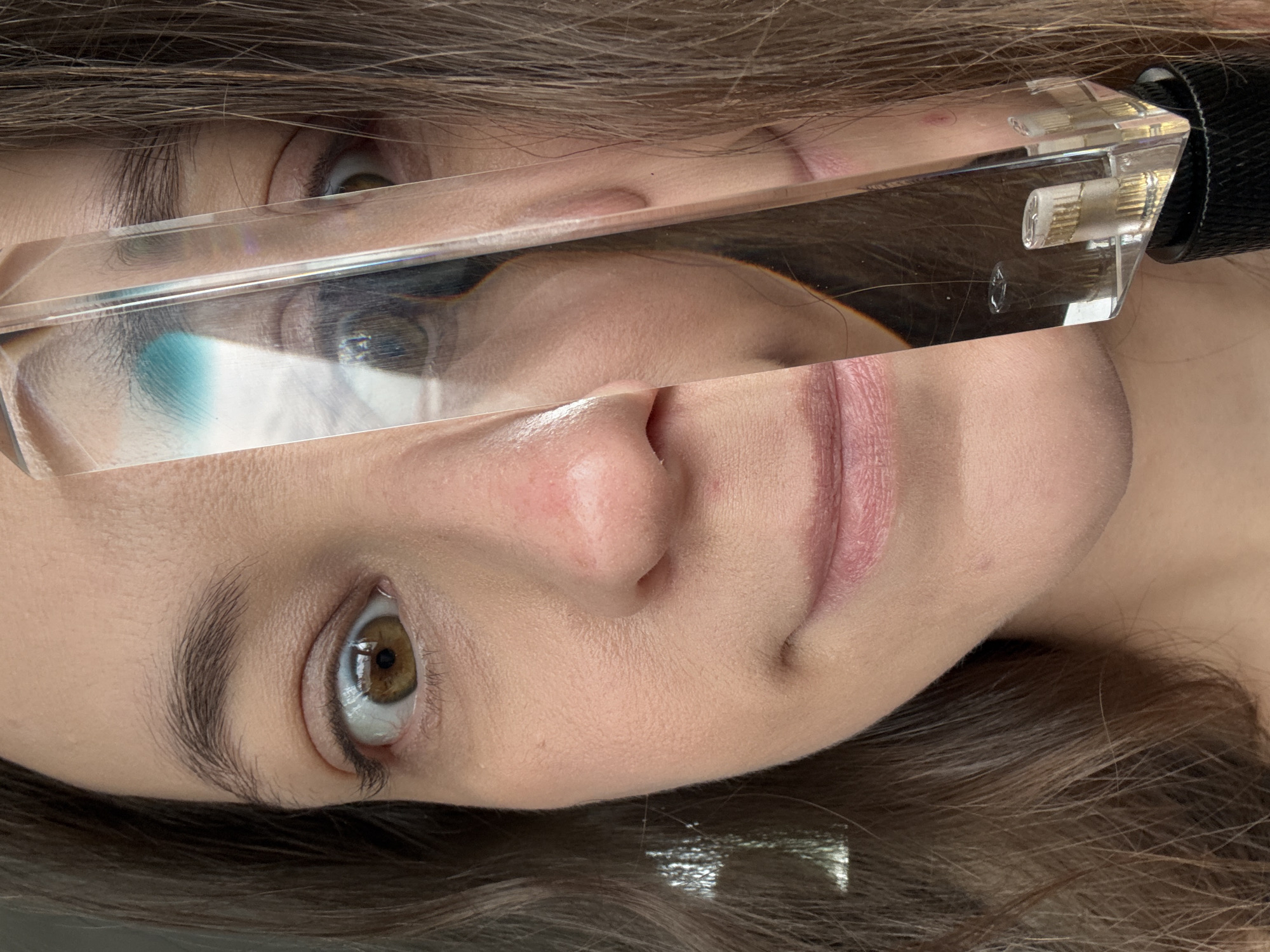 A medium close-up, slightly rotated portrait shot depicts a person's face bisected by a clear, rectangular prism. The individual has light skin, brown hair, and brown eyes. The prism distorts and refracts the upper portion of the face, particularly the left eye, creating a warped reflection. The nose and mouth are visible in the lower portion of the face. The right side of the prism has a black handle or attachment visible. No contextual information is provided that points to the location of Chaikovsky, Russia.