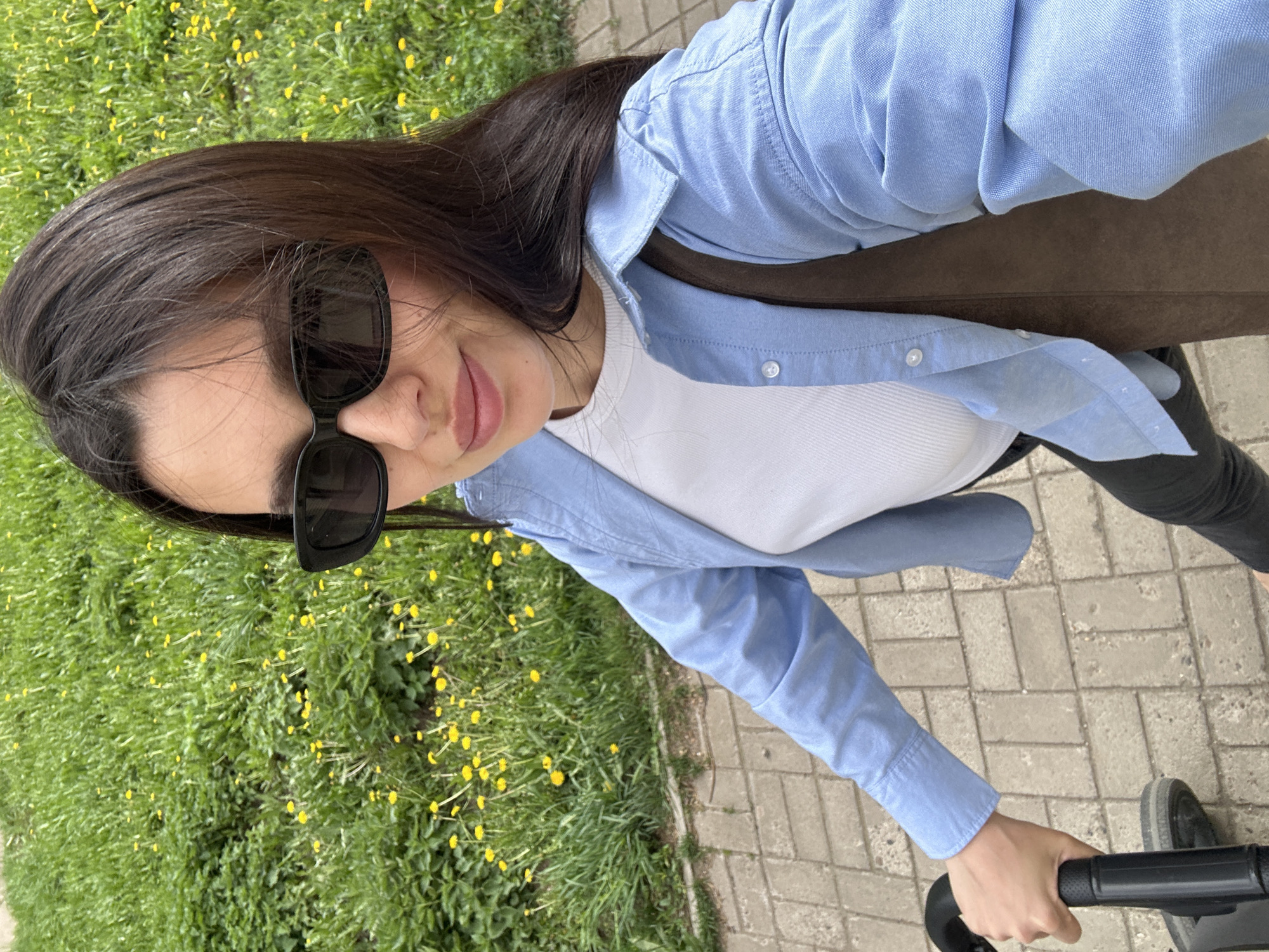 A woman is seen from an elevated angle, appearing to be in the city of Chaikovsky, Russia, based on visible elements characteristic of that location. She is wearing dark sunglasses, a light blue long-sleeved shirt over a white ribbed top, and dark pants. She is holding the handle of a stroller, suggesting she is either walking with it or preparing to. The background shows green grass with small yellow flowers, and a paved walkway. The scene suggests an outdoor setting during daylight hours.
