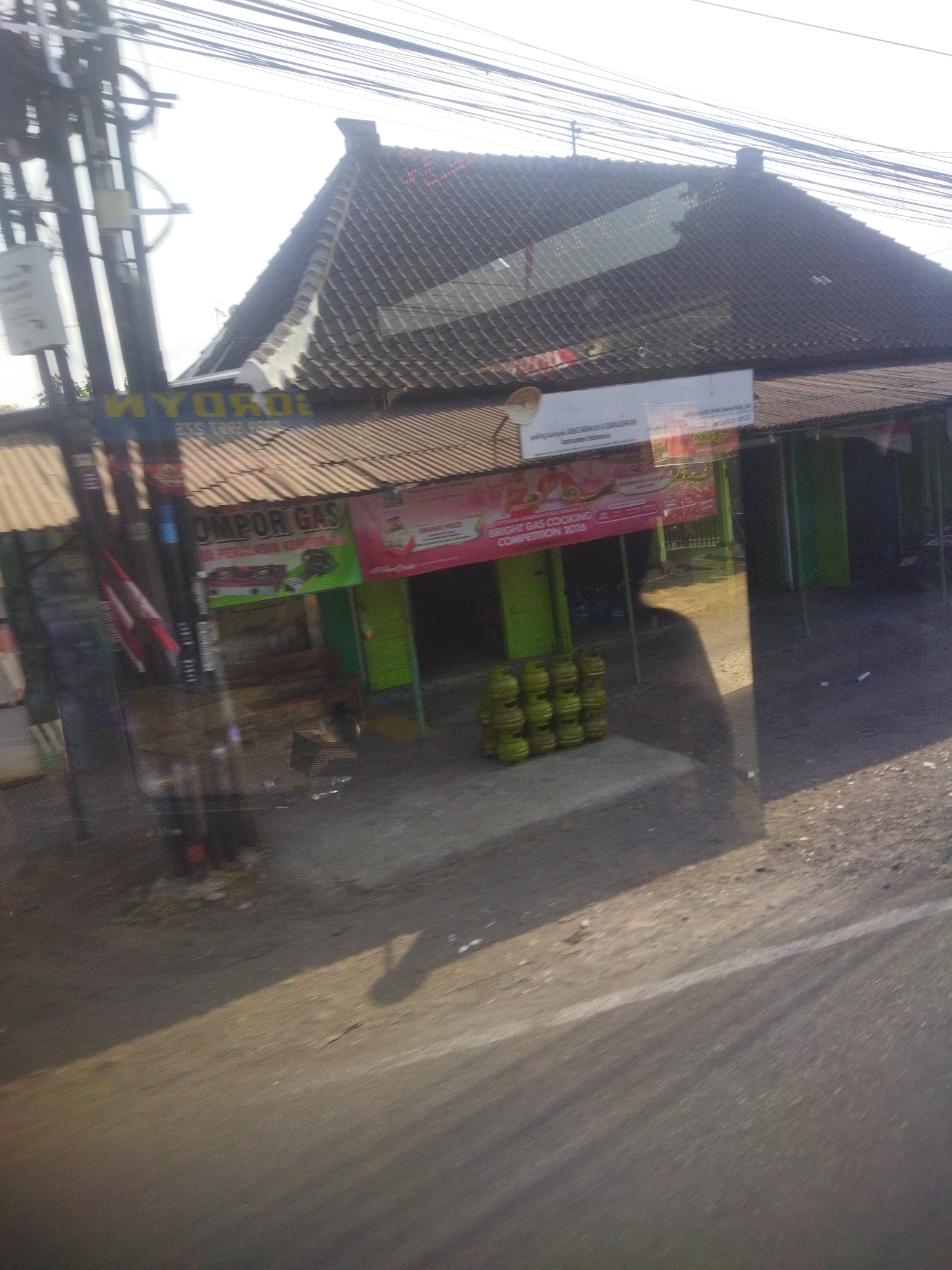The image depicts a roadside scene in Nguter, Indonesia, at what appears to be a commercial establishment. A tiled roof structure with a prominent chimney stands out against a bright, overcast sky. The building's facade features a corrugated metal awning supported by poles. Visible signage includes the words "MYOKOR" and "OMPOR GAS," suggesting a gas supply business or a store selling gas-related products. Piles of green gas cylinders are stacked on the ground in front of the building. The foreground shows a paved road, indicating a street-level view, likely captured from a moving vehicle due to the slight motion blur. Shadows are cast across the scene, indicating daylight. The environment appears somewhat rural or suburban, with overhead power lines and a general lack of dense urban infrastructure.