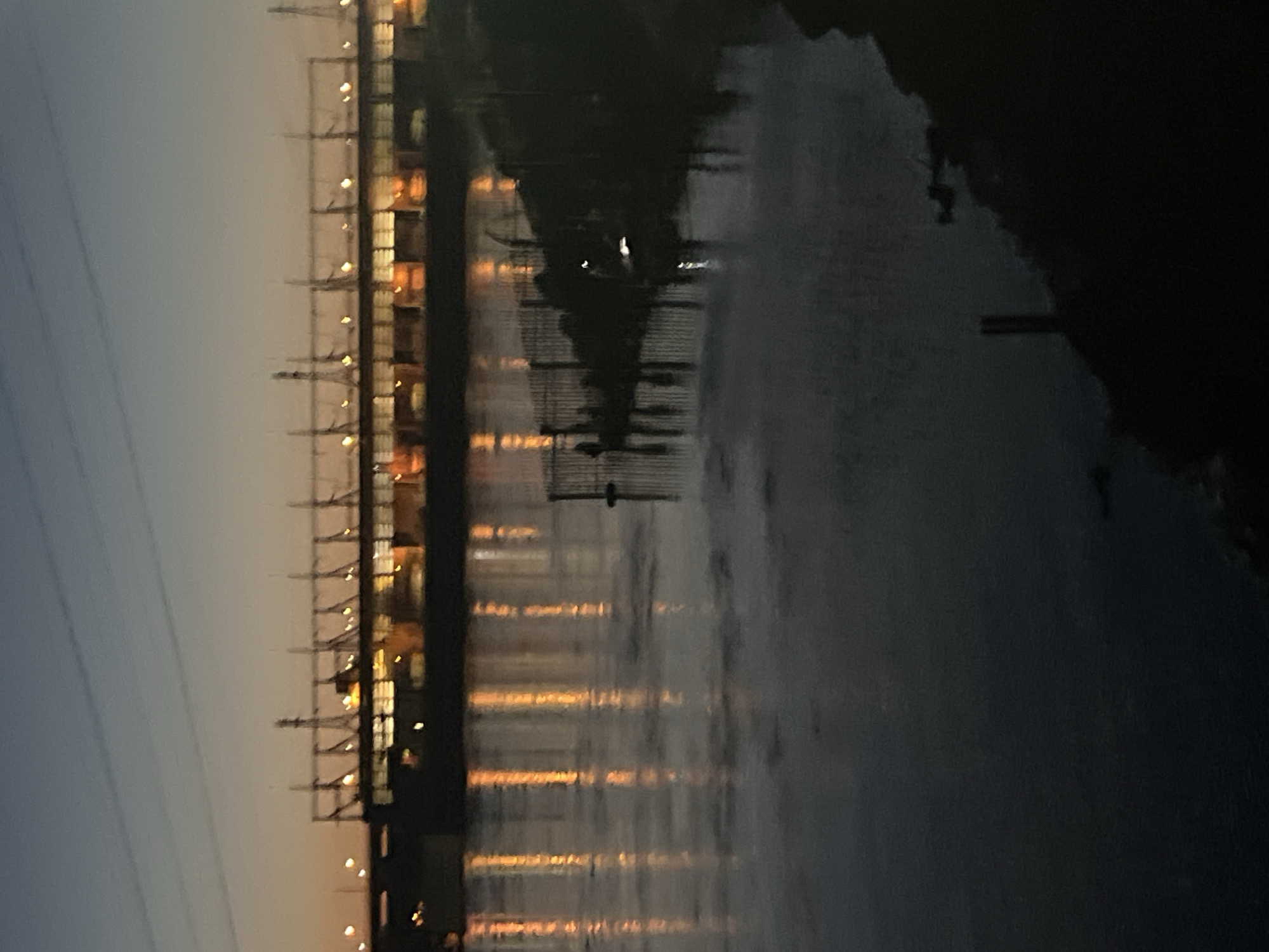The image captures a waterfront scene at dusk in Chaikovsky, Russia. A large dam or hydroelectric power station is prominently featured in the background, its structures silhouetted against the fading sky. Lights from the facility illuminate the water, creating reflections that streak across the surface. The foreground consists of a body of water, likely a river or reservoir, with a dark embankment visible on the right side. A section of fencing or a railing runs along the edge of the water in the mid-ground, casting shadows. The overall atmosphere is serene and slightly industrial, typical of a city with significant infrastructure.