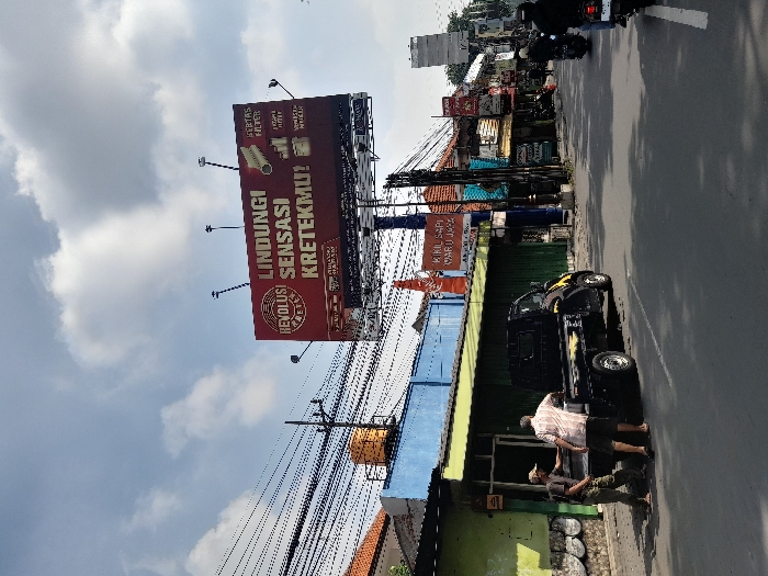 The image shows a street scene in Kecamatan Waru, Indonesia, located at coordinates -7.352 latitude and 112.729 longitude. A large billboard displays an advertisement in Indonesian for a product called "Krektek." Two men in short-sleeved shirts stand in the street near a black pickup truck parked on the side of the road. Power lines run across the scene, and other buildings can be seen in the background. In the distance, motorcycles are traveling on the road. The sky is partly cloudy.