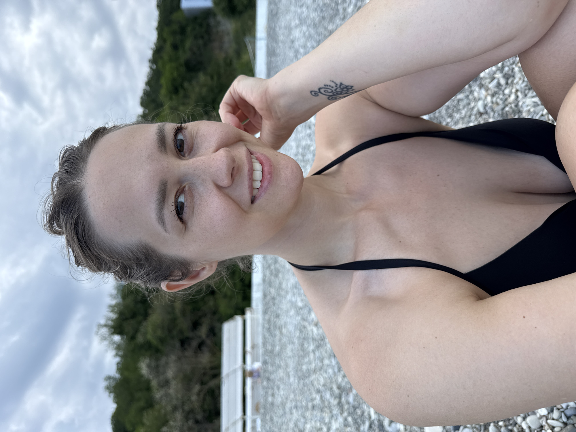 A woman is taking a selfie on a pebble beach. She is wearing a black bikini and has her hair tied up. She is smiling and resting her hand on her cheek. There is a tattoo on her left forearm. In the background, there is a cloudy sky, lush green trees, and some white structures that appear to be beach umbrellas or shelters. The location is Nebug, Russia.