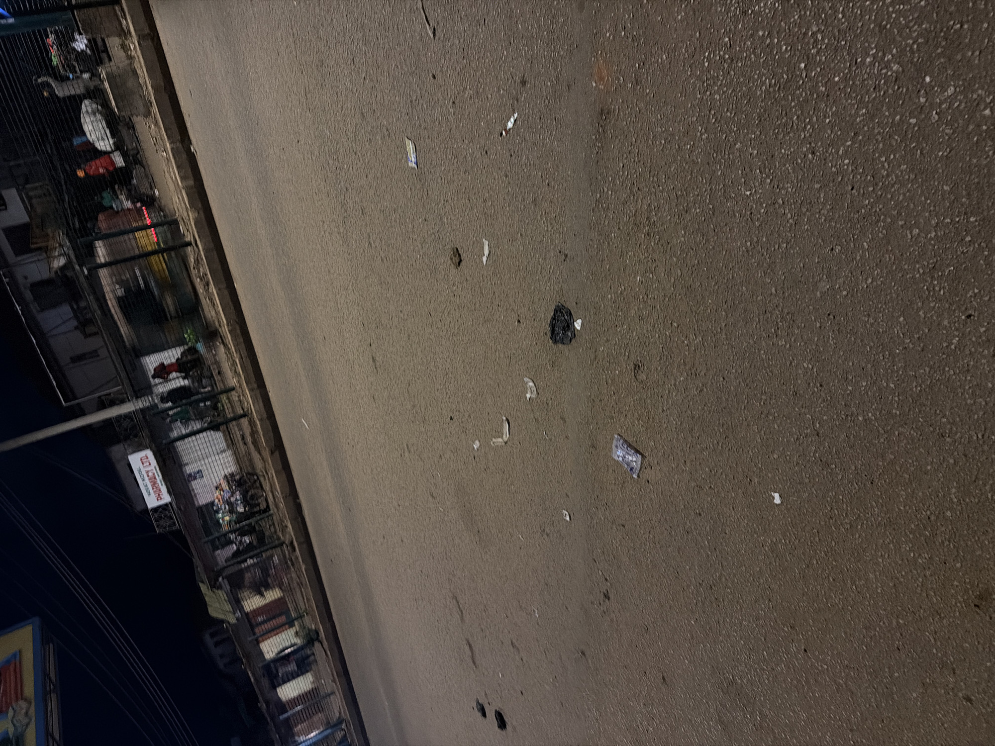 The image depicts a street scene at night in Bukuru, Nigeria, with a focus on the road surface which is littered with various debris. In the background, several small shops are visible, some with open fronts and others with shutters. A sign for "PHARMACY LTD." is prominently displayed on one of the buildings. Several individuals are present, some standing near the shops and others appearing to be walking or engaged in activity. A motor vehicle, possibly a motorcycle, is parked in front of one of the shops. The road itself is asphalt and shows signs of wear and tear, with scattered pieces of paper, plastic wrappers, and a black plastic bag. The overall lighting suggests it is after sunset, with streetlights or building lights illuminating the area.