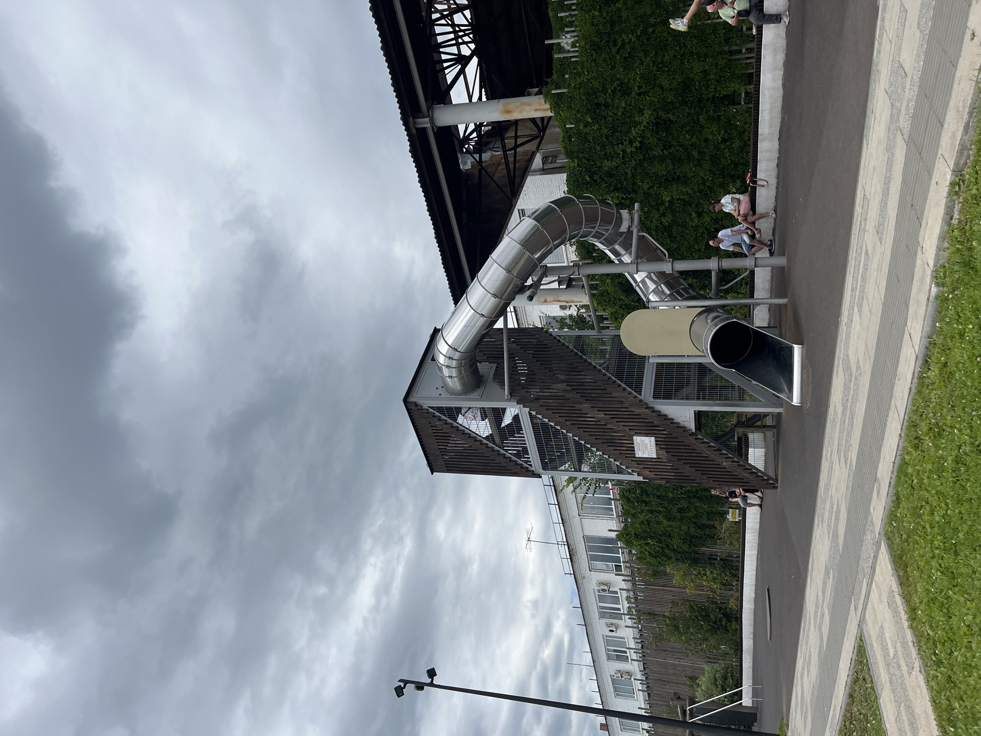 The image depicts an outdoor playground with a large, silver metal slide as the central feature. The slide is integrated into a wooden and metal structure with stairs and railings. In the background, there is a long, white industrial-looking building with many windows. To the right of the building, a darker, dilapidated structure with a corrugated roof is visible. A dense green hedge runs along the fence line separating the playground from the buildings.
Several people are present in the scene. Two individuals are sitting on a bench in front of the hedge, appearing to be conversing. Another person is seated on a ledge to the right, with their legs bent. A person is partially visible at the top of the slide structure. A lamp post with multiple lights stands to the left of the playground. The ground is a dark, rubberized surface, with a paved walkway and grass in the foreground. The sky is overcast with heavy, gray clouds.
The image was taken on May 25, 2023, at 16:40:05 in Saratov, Russia.