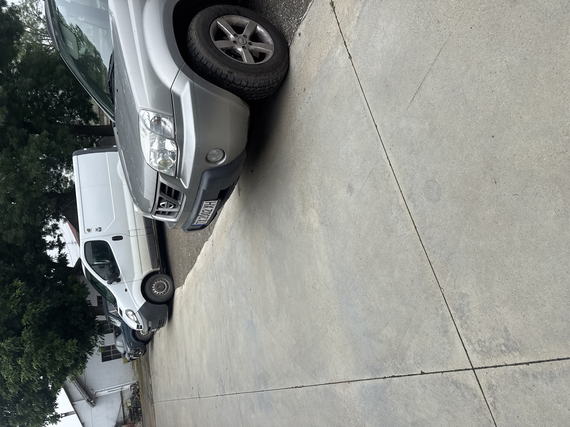 The image, originally oriented vertically, captures a static outdoor scene in Sarnevo, Bulgaria, featuring several parked vehicles on a concrete surface. In the foreground, a light grey SUV, appearing to be a Nissan X-Trail, is prominent, facing towards the right side of the frame. Its front bumper, headlight, grille, and a portion of its front wheel are clearly visible. The license plate on this SUV reads "TX 2002 XH," with "TX" being the identifier for the Veliko Tarnovo region in Bulgaria, confirming the location context.
Behind the grey SUV, a white cargo van, possibly a Renault Trafic or similar model, is parked, with its front and side clearly visible. Further in the background, another dark-colored vehicle is partially visible behind the white van. The ground is a light grey concrete pavement, showing distinct expansion joints that form lines across the surface.
To the left and in the background, dense green trees are visible, providing a natural backdrop. Partially obscured by the trees and the white van are white-painted buildings, one with a visible window, suggesting a residential or light industrial area. The lighting is soft and diffused, indicating an overcast day or possibly early morning/late afternoon, as there are no harsh shadows. No specific human activity or event is taking place; the scene depicts parked vehicles in their environment.