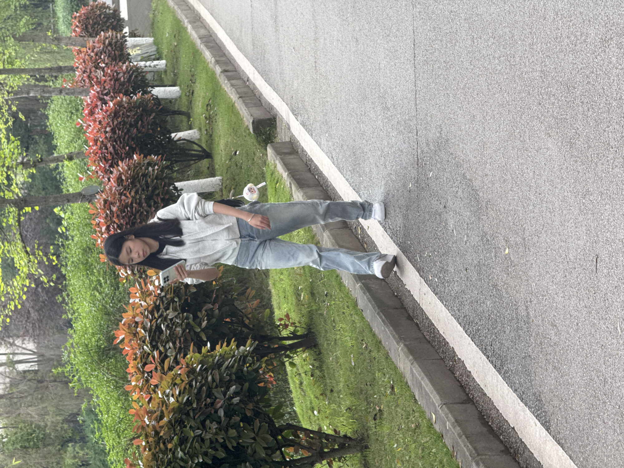 The image depicts an individual standing on the side of a roadway. The person is facing to the left side of the frame and is holding a mobile device in their hands. A row of bushes lines the edge of the roadway. The location is Jiangjin, China.