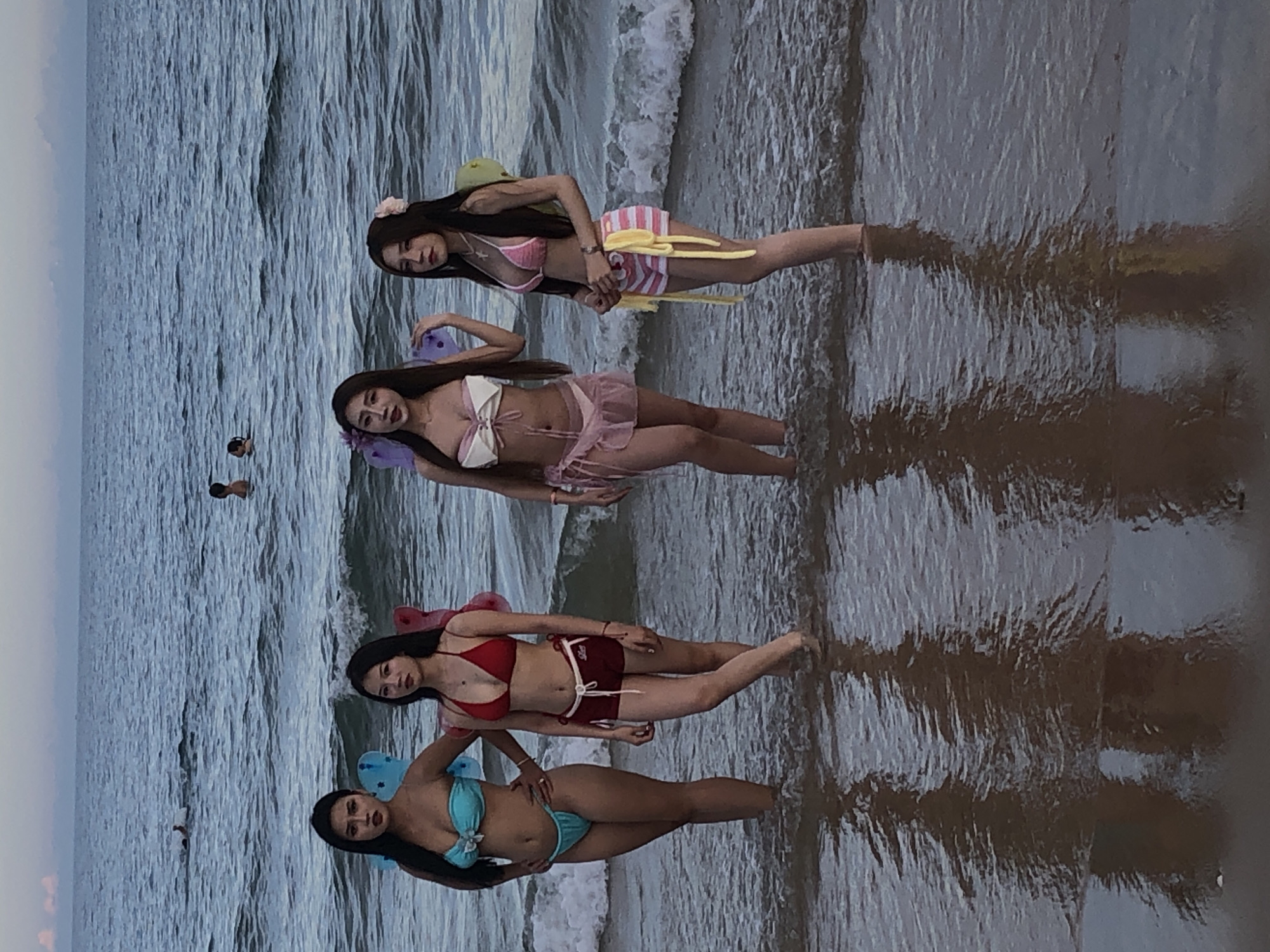 Four young women stand in shallow ocean water on a beach. They are positioned from left to right in the frame. All are wearing bikinis and have fairy wings attached to their backs. The woman on the far left wears a teal bikini. The second woman from the left wears a red bikini top and maroon shorts. The third woman from the left wears a white bikini top with a pink trim and a sheer pink skirt. The woman on the far right wears a pink bikini top with white stripes and matching pink and white striped shorts with yellow ties. The water is calm with small waves. In the background, two individuals can be seen in the water further out, and the sky is a soft gradient of pink and blue. The reflections of the women are visible in the water. The setting appears to be a beach in Phu Thuy Ward, Vietnam.