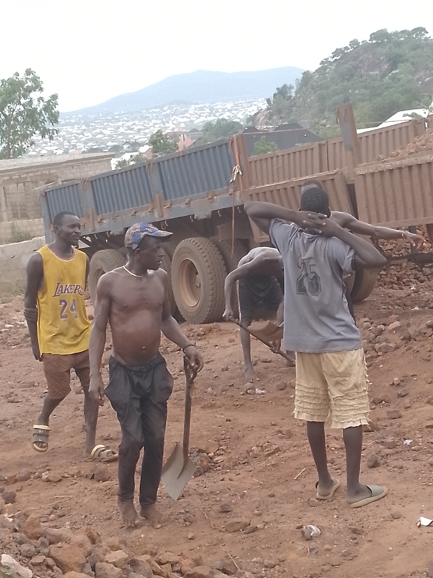 The image depicts four men in a dusty, rocky construction or excavation site in the city of Unknown, Unknown. A large dump truck with multiple axles is in the background. One man, wearing a yellow basketball jersey with the number "24", brown shorts, and sandals, is walking to the left. Another man, shirtless with a cap and dark pants, is holding a shovel. Another man is shirtless, using a shovel. The fourth man is wearing a grey shirt with the number "25" on it and light-colored shorts. The terrain is dirt and rocks. Buildings and a distant mountain are visible in the background.