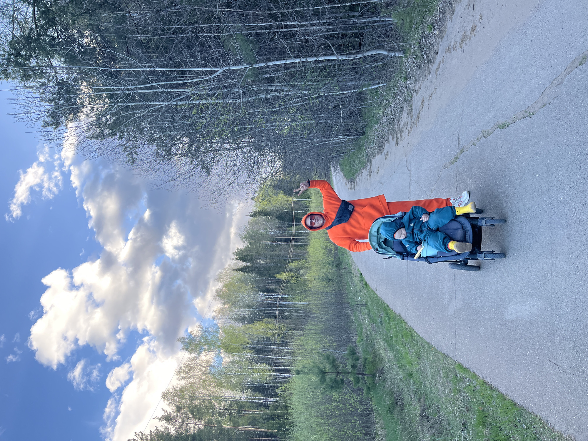 A man in an orange hoodie and sunglasses is on a paved path with a stroller containing a small child wearing a blue outfit and yellow boots. The path is flanked by a green grassy verge on one side and a dense forest of trees on the other. A partly cloudy blue sky is visible through the trees. The location appears to be in a park area. Although it is difficult to identify the exact location with certainty, given the information, the scene is likely in or near the city of Chaikovsky, Russia.