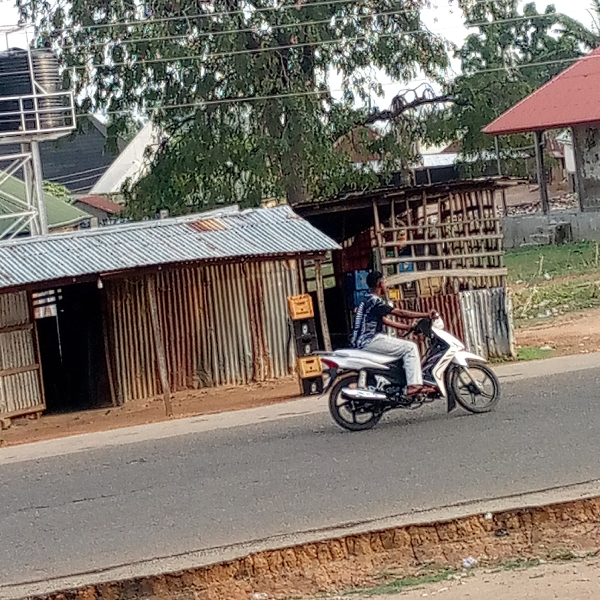 An eye-level shot captures a person riding a white motorcycle on a paved road, likely in a rural or semi-urban setting. The rider, wearing a patterned shirt and light-colored pants, is positioned with their back mostly to the viewer, facing right. The motorcycle is white with black accents and wheels.
To the left of the rider, there is a building or stall with walls made of rusty, vertical corrugated iron sheets and a matching corrugated roof. An open doorway reveals a dark interior. Next to this structure, a stack of yellow and dark-colored plastic crates, possibly for beverages, is visible. Behind the motorcycle, there's another wooden, open-sided structure or stall, also seemingly made with corrugated iron or wooden planks, suggesting a marketplace or roadside shop.
The road itself is asphalt, with a light brown dirt or grass shoulder to the right. In the background, lush green trees dominate the upper half of the frame, with power lines crisscrossing through them. Several other buildings with various roof types (including green, white, and a distinct red-tiled roof on the far right) are partially visible through the foliage and above the foreground structures. One structure on the far left has a metallic framework, possibly supporting a water tank.
The lighting suggests it is daytime, with soft shadows indicating a bright but possibly overcast sky or late morning/early afternoon. No specific text is clearly legible on the crates or buildings, though some symbols or logos are faintly visible on the yellow crates (appearing like an 'H' or 'I-I' shape). The overall scene depicts a mundane moment of daily life in an outdoor environment.
