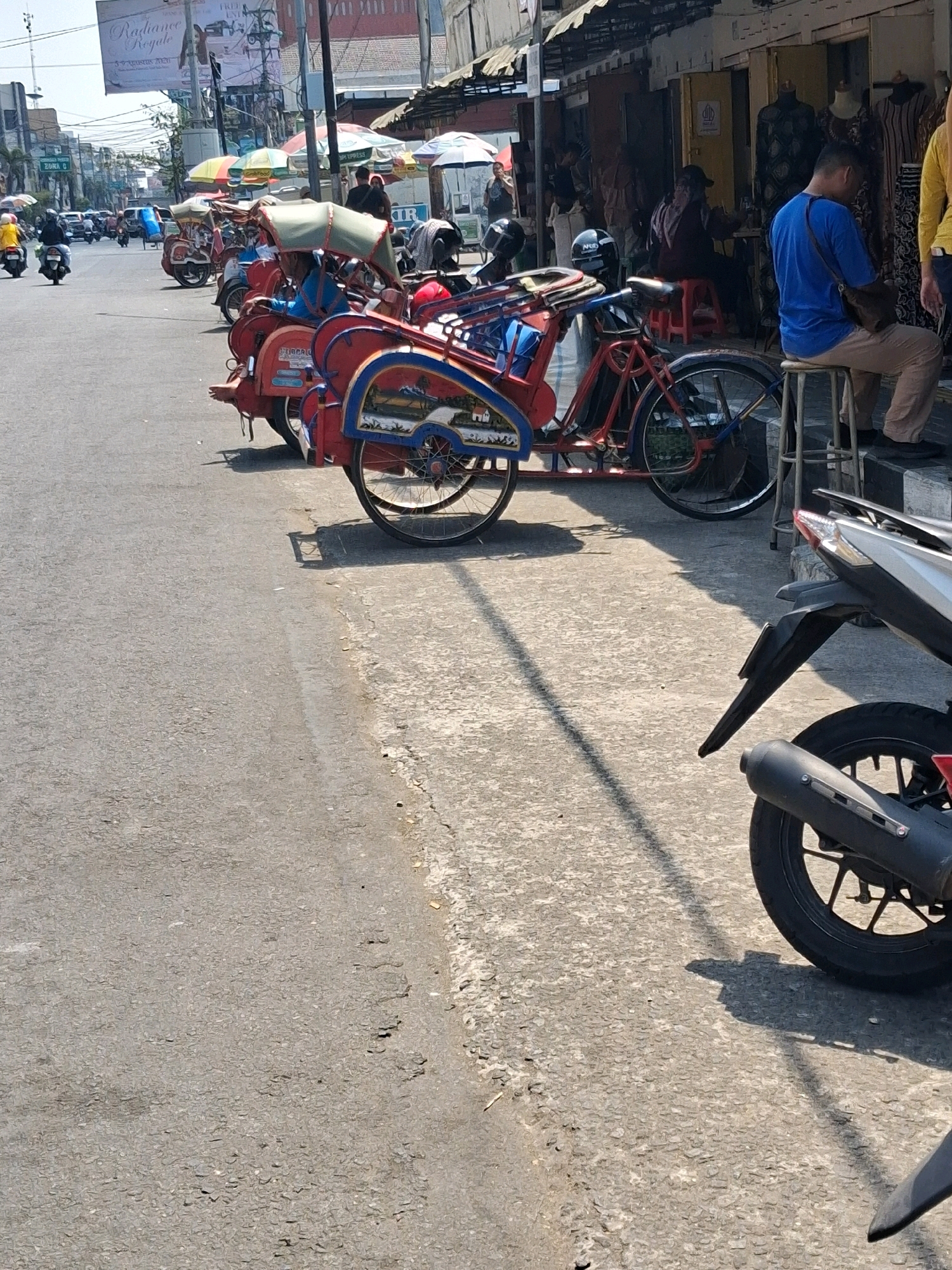 The image depicts a street scene in Surakarta, Indonesia, likely during daylight hours given the bright sunlight and shadows. The foreground is dominated by the rear sections of two motorcycles, one silver and black, the other predominantly black with a red accent. In the midground, several brightly colored pedicabs, known locally as "becak," are parked along the side of the road. These becaks are red and blue with decorative panels and fabric awnings.
Further back, on the right side of the image, is a row of shops with open doorways. A man wearing a blue t-shirt and light-colored pants is seated on a stool in front of one of the shops, looking downwards. Another person, partially obscured, is also seated nearby. Umbrellas of various colors are visible over some of the shops, suggesting a market or commercial area.
The street itself is paved with asphalt, and a concrete sidewalk runs along the right edge of the road. The overall impression is of a bustling urban environment, possibly a commercial district where local transportation like becaks and motorcycles are common. The presence of clothing displays in the shop windows indicates a retail setting.
