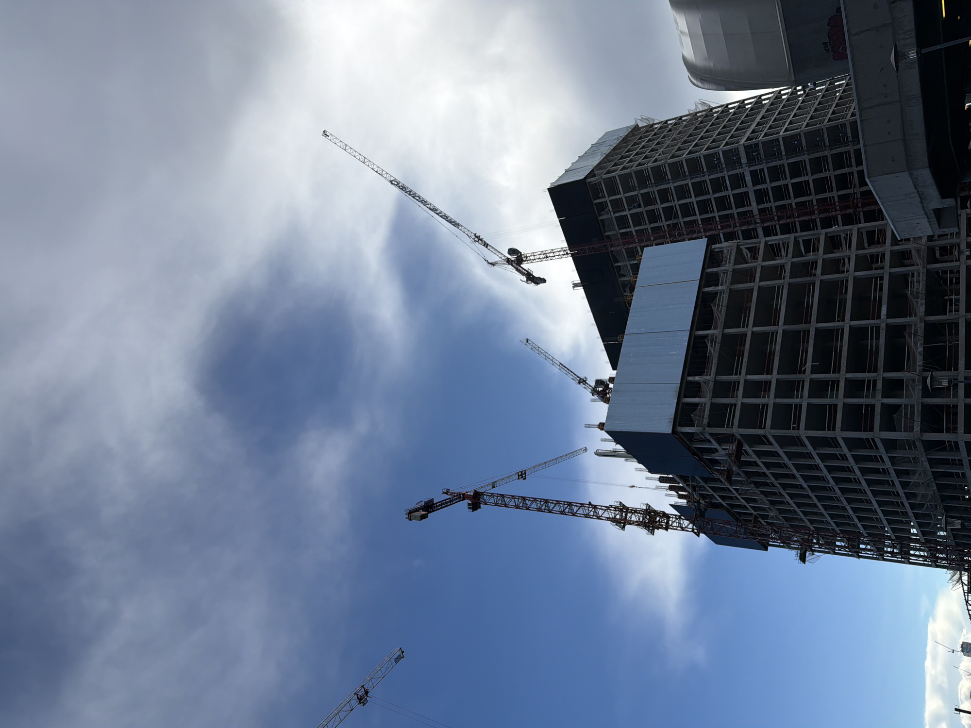 The image shows a construction site with several tower cranes against a bright, partly cloudy sky. The main subjects are two large, unfinished buildings. One building is a towering structure with a visible concrete framework and some metallic cladding. The other building appears to be of similar scale, also in a state of construction.
Multiple tower cranes are active on the site. One large crane with a red and grey boom is positioned prominently, its arm extended over the buildings. Other cranes, with lighter colored booms, are visible in the distance and closer to the foreground, also appearing to be in operation. The angle of the shot is from below, looking up at the construction and the sky.
The lighting suggests it is daytime, likely mid-morning or afternoon, given the brightness of the sky and the shadows cast. The clouds are wispy and scattered, indicating fair weather conditions. There are no people visible in the frame, but the presence of the active cranes strongly suggests ongoing construction work. The scene is set in an urban environment, as evidenced by the scale of the buildings and the machinery. No text is clearly discernible in the image.