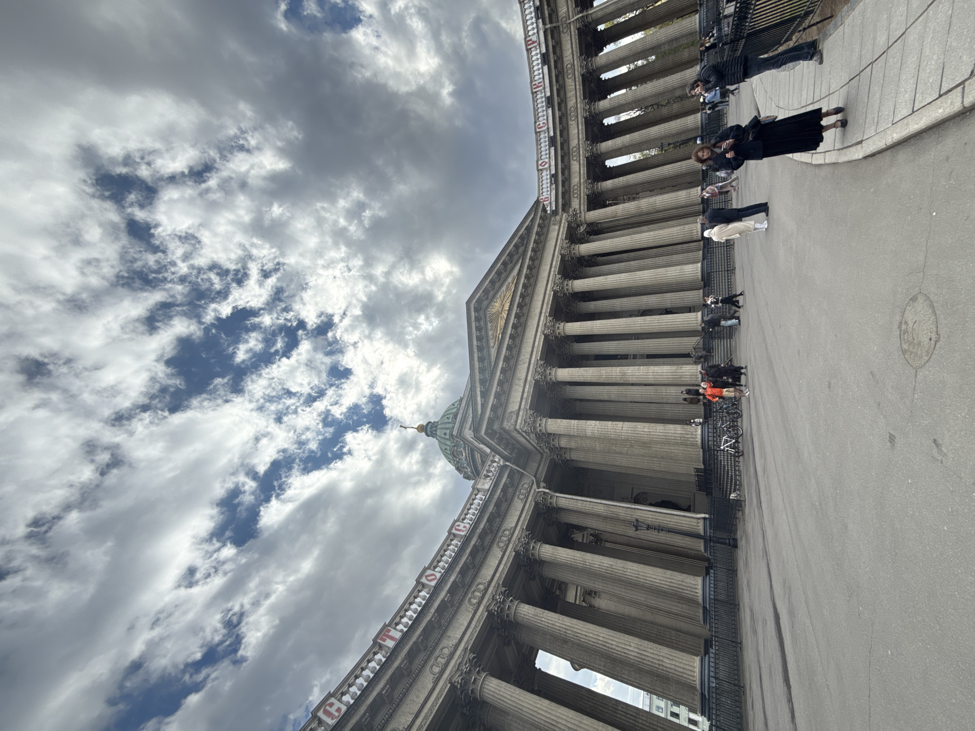The image captures a view of the Kazan Cathedral in Saint Petersburg, Russia. The cathedral's facade, distinguished by its neoclassical architectural style and numerous columns, is prominently displayed against a backdrop of a cloudy sky with patches of blue. People are seen walking and standing along the sidewalk adjacent to the cathedral. Bicycles are parked near the columns. The perspective is from below, tilting upwards toward the cathedral's dome and the sky, giving a sense of its grandeur and scale.