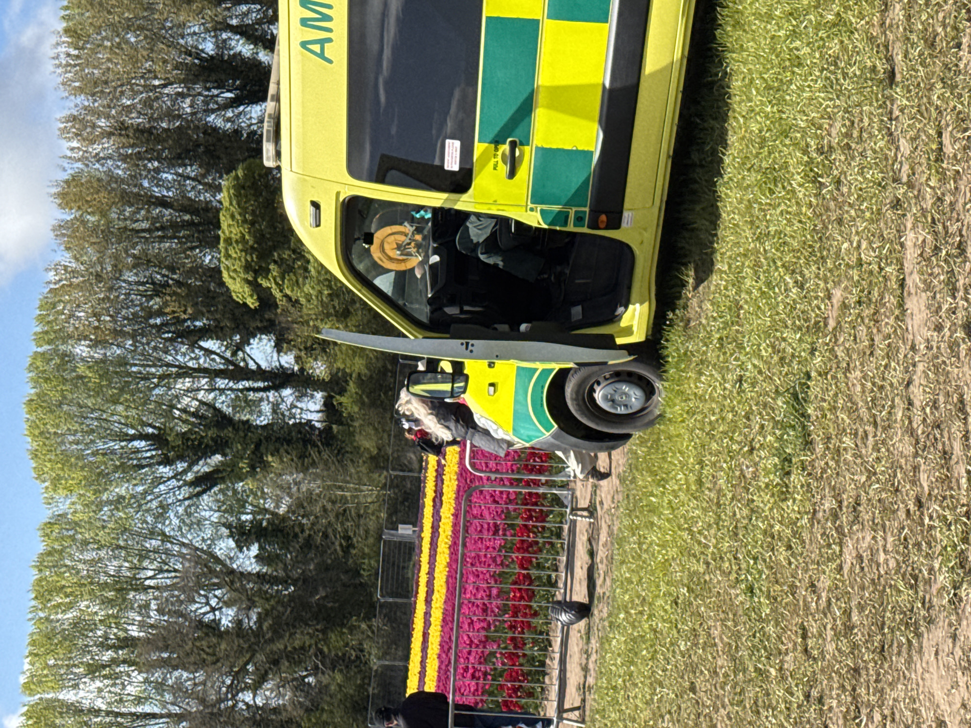 The rotated image captures a vibrant daytime scene featuring a bright yellow ambulance parked beside a colorful tulip field under a partly cloudy sky. The ambulance, a UK model with distinctive yellow and green Battenburg markings, has its driver's side door open. A person, possibly in uniform, is visible in the driver's seat, and a hat or helmet is on the dashboard. The word "AMBULAN" is visible on the vehicle's side, and "PULL TO OPEN" is printed near the door handle.
Adjacent to the ambulance is a meticulously arranged field of tulips, showcasing alternating rows of vivid yellow and deep red/magenta flowers. Metal crowd control barriers separate the vehicle and onlookers from the cultivated floral display. Several people are present, including one individual with light-colored hair near the front of the ambulance and another person in dark clothing viewing the flowers from behind the barriers.
In the background, a dense line of trees with abundant green foliage stands against the clear sky, suggesting a rural or semi-rural setting. The ground immediately surrounding the ambulance on the right side appears as rough grass or earth. The scene is well-lit and tranquil, indicating pleasant weather conditions. No emergency activity is depicted; the ambulance seems to be parked, possibly on standby or at an event, in the King's Lynn and West Norfolk area.