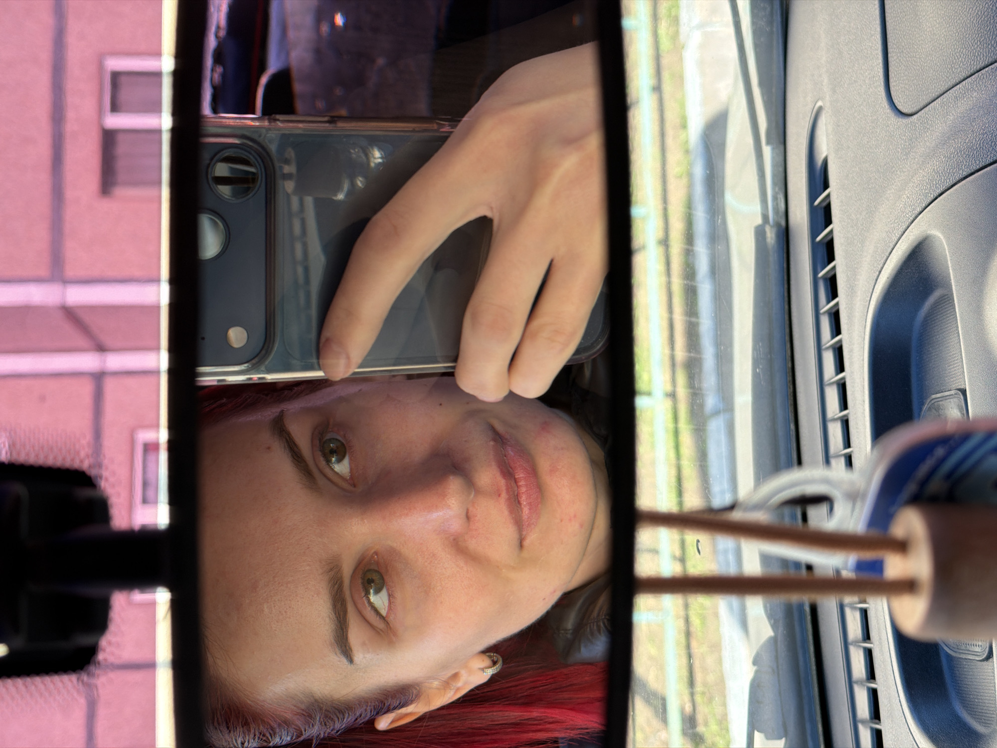 A selfie is captured through a car's rearview mirror, featuring a person with reddish hair and green/hazel eyes. The subject's face is reflected sideways in the mirror, displaying a neutral expression. They are holding a smartphone, visible with its dual camera lenses and a clear case, which is being used to take the photograph. A silver hoop earring is visible in their left ear.
Around the mirror, parts of the car's interior are visible, including a textured gray dashboard or door panel with an air vent. To the right of the mirror, a car air freshener with two wooden sticks is present.
The reflections in the mirror show the surroundings: on the left, a distinct pink or reddish building with windows suggests an urban or residential area. On the right, green foliage or grass, possibly alongside a fence, indicates an outdoor setting. The bright, clear lighting confirms it is daytime. This scene is set in Chaikovsky, Russia. No text is visible in the image.
