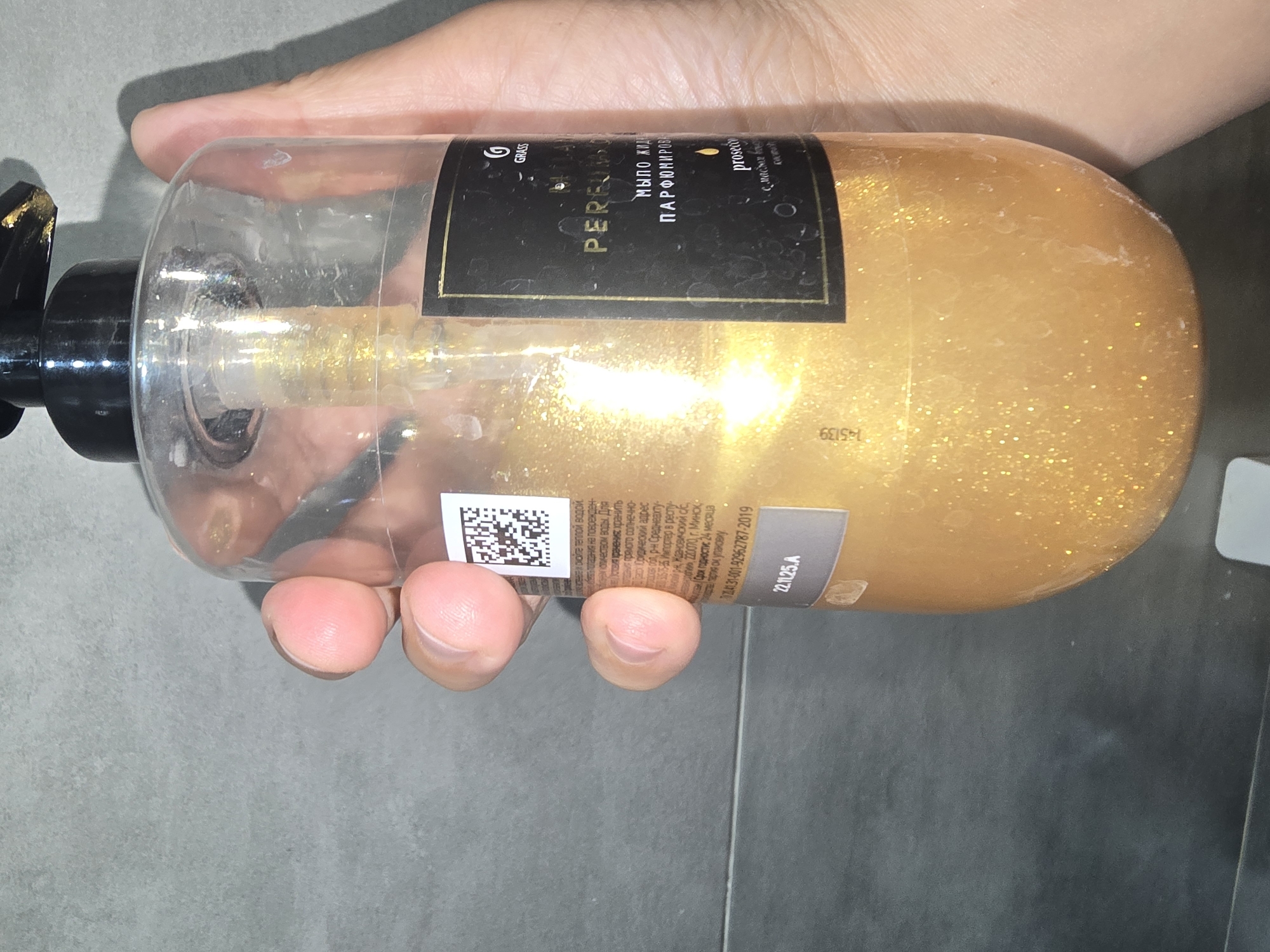 A person's hand is holding a clear plastic bottle of golden, glittery liquid soap with a black pump dispenser. The bottle has a black label with gold text. The background appears to be a tiled wall, possibly in a bathroom. The image does not contain any information that would allow for its location to be identified as Ulan-Ude, Russia, or any other specific city.