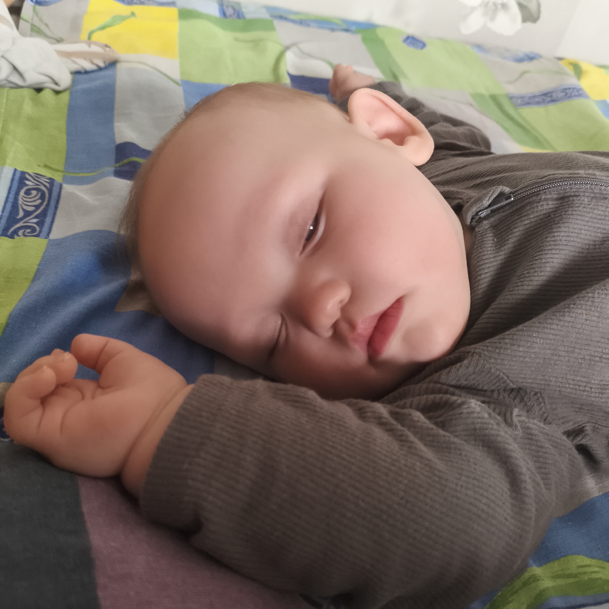 A baby is sleeping on a bed covered with a colorful patchwork quilt, featuring blue, green, and yellow squares. The baby is lying on their right side, wearing a grey corduroy-like garment with a visible zipper near the neck. The baby's right arm is extended on the quilt, with their fingers curled. The location is Chaikovsky, Russia.
