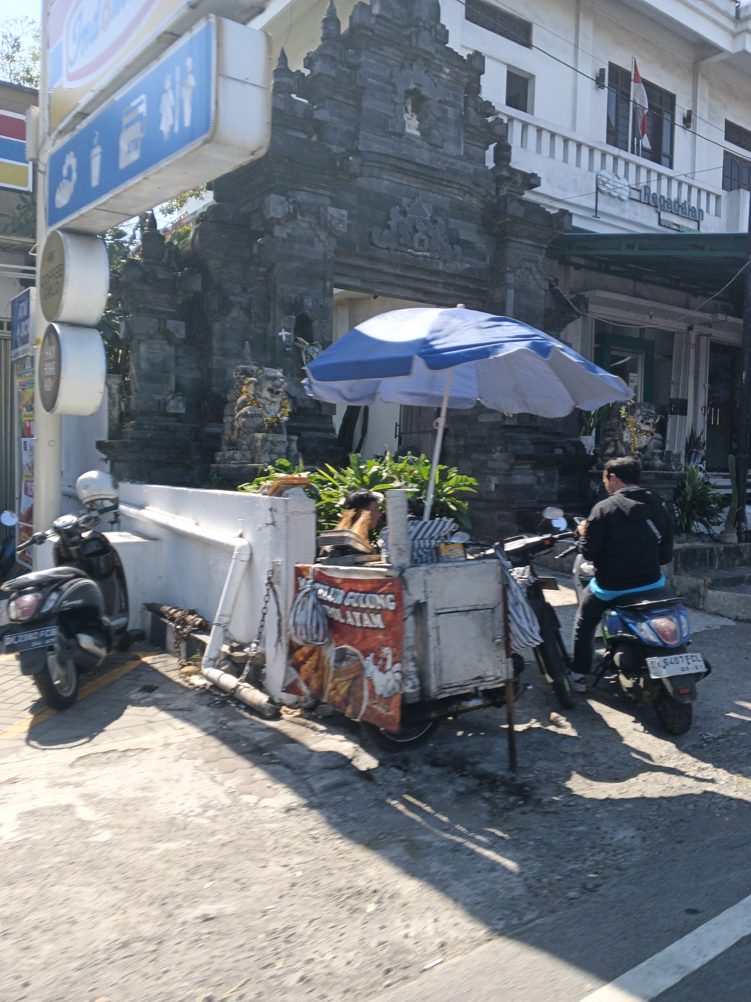 In Abiansemal, Indonesia, a street scene unfolds under a bright sun, casting distinct shadows on the asphalt. A man, facing away from the camera, sits on a blue scooter, his back to a food cart. He wears a dark jacket over a bright blue shirt. The food cart itself is weathered, displaying an advertisement with images of food. To the left of the cart, a black scooter is parked. In the background, a traditional stone structure, possibly an entrance to a temple or historical site, stands prominently. The structure is adorned with intricate carvings and a lion statue. Above the stone structure, a white building with a balcony and a red and white flag visible, suggests commercial or residential space. A blue and white umbrella shades the food cart. The environment appears to be a bustling street, indicated by the presence of multiple vehicles and signs, one of which, on the left, features blue icons for drinks and food, and figures of people.