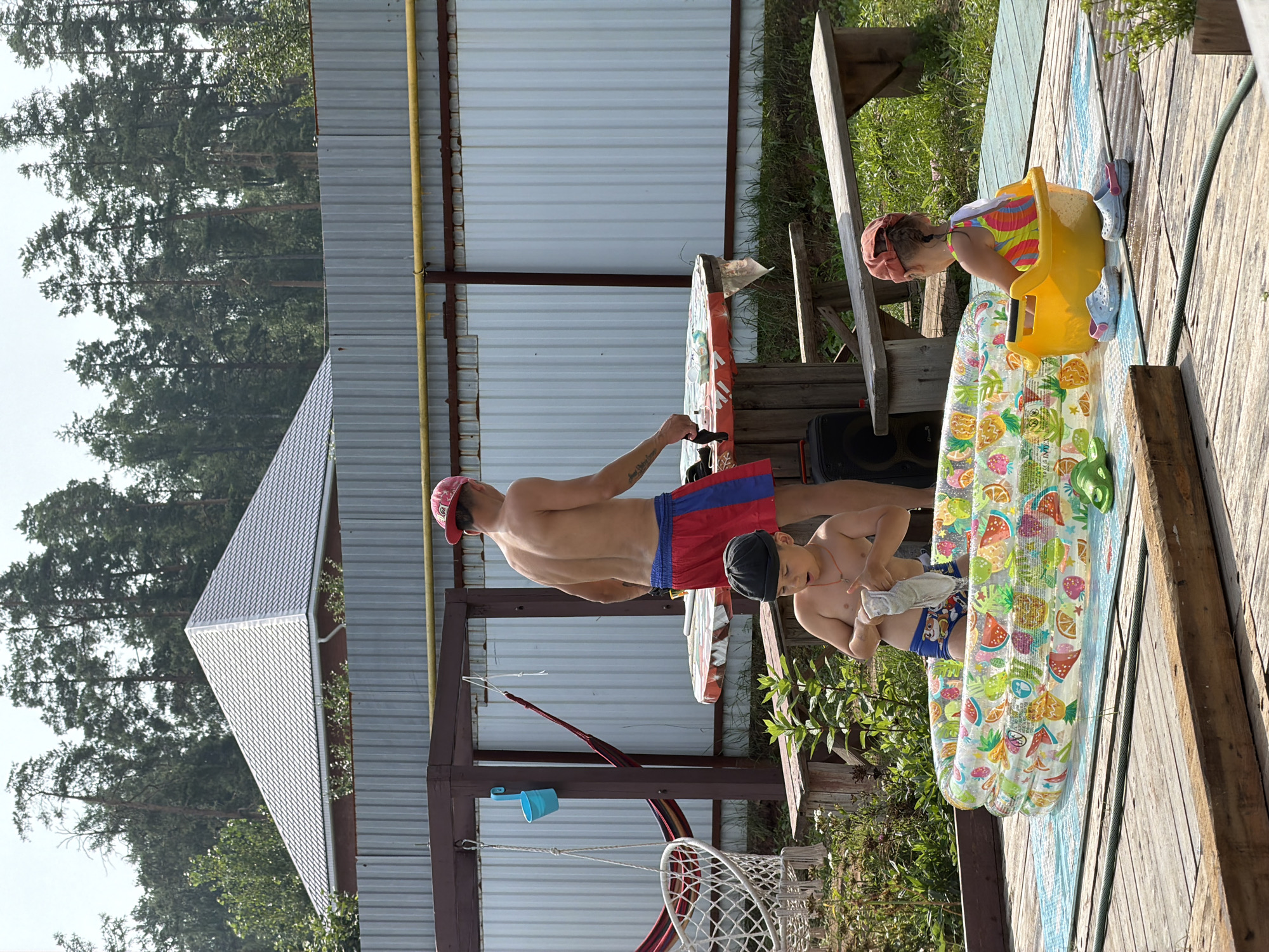 In a backyard setting, likely in Chaikovsky, Russia, an adult male wearing a red baseball cap and red and blue swim shorts stands near a round table covered with a red and white checkered tablecloth. He appears to be holding a small black object in his right hand. To his left, a young boy, also shirtless and wearing blue swim trunks with a pattern, stands in an inflatable pool filled with water and decorated with fruit images. The boy is holding a white cloth. To the right of the inflatable pool, a young girl in a colorful swimsuit is seated in a yellow plastic tub. Various outdoor furniture, including a hammock, a white woven chair, wooden benches, and a picnic table, are visible in the background. The area is surrounded by a corrugated metal fence and tall pine trees. The ground is a wooden deck, with grass and plants visible in the periphery. The overall scene suggests a warm day, with recreational activities for children taking place outdoors.