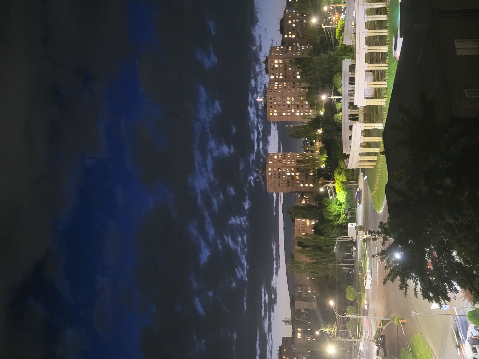The image depicts a night scene in Yerevan, Armenia. High-rise apartment buildings with illuminated windows are visible along with streetlights illuminating the roadway and sidewalks. Trees line the streets. A colonnade structure is present. The sky is a dark blue with scattered clouds. Several cars are present on the roadways.